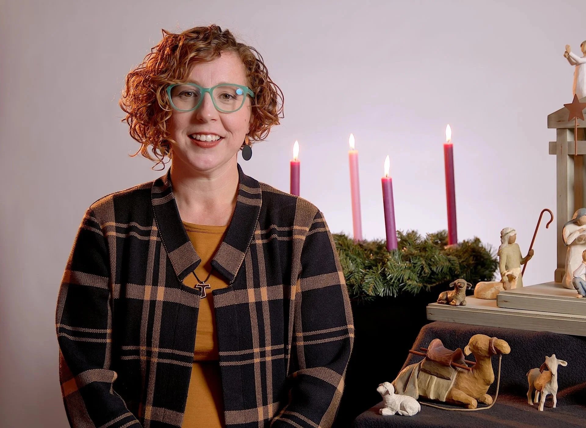 A woman with curly red hair, green glasses, and a checkered jacket sitting in front of Christmas decorations including a wreath with pink candles and nativity figurines.
