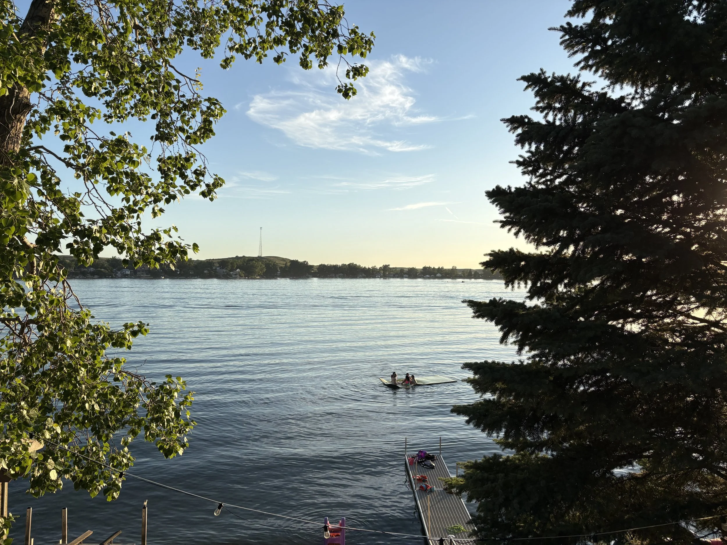 A small lake with the other shore in sight on the horizon, a blue sky with wisps of white cloud, the setting sun shining across the lake, with a poplar and an evergreen in the foreground on each side of the photo. A handful of kids play off a dock.
