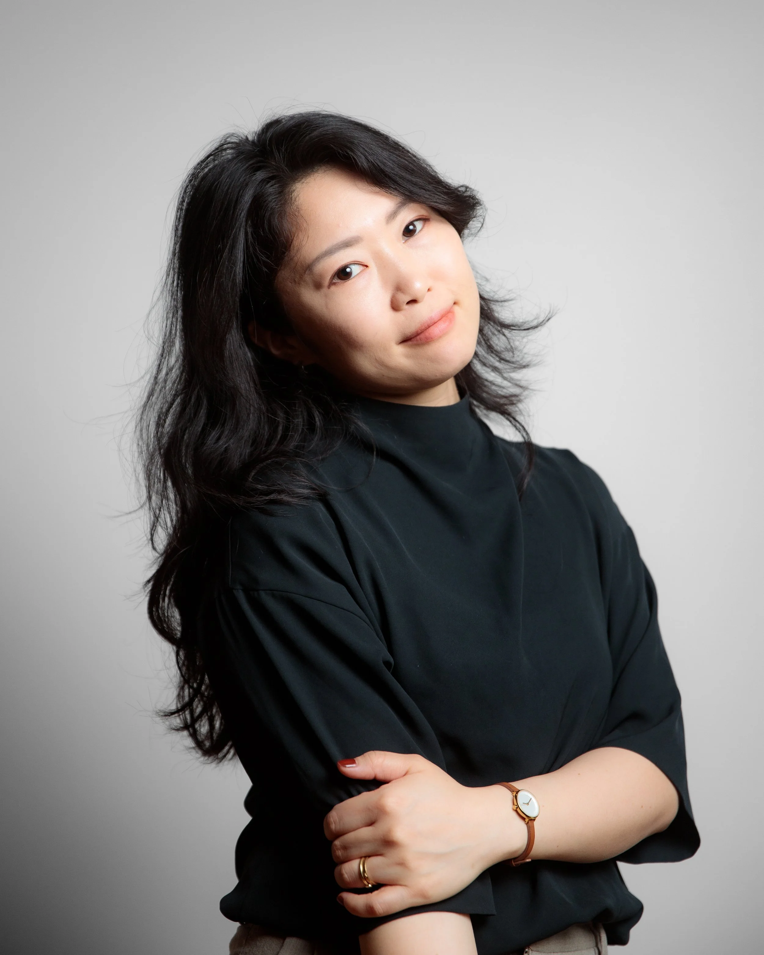 A woman with dark, wavy hair and light skin, wearing a black top and a gold wristwatch, standing against a plain gray background. She has a gentle smile and is crossing her arms.