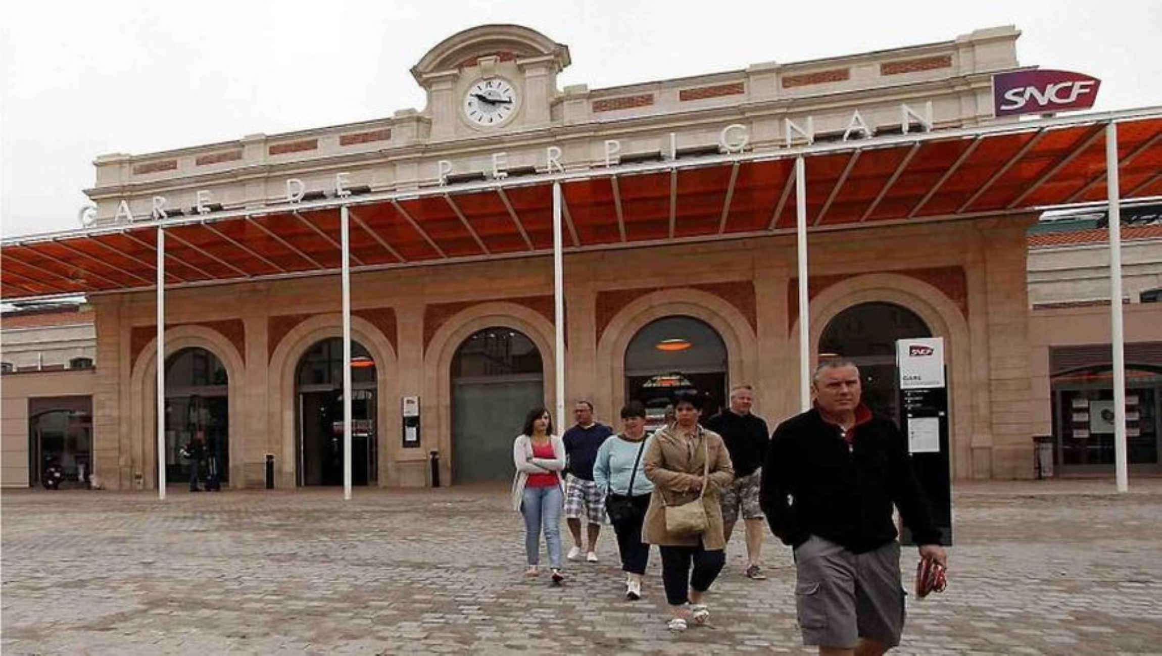 Gens marchant devant la gare Gare de Perpignan, en façade de style classique avec horloge et enseignes SNCF.