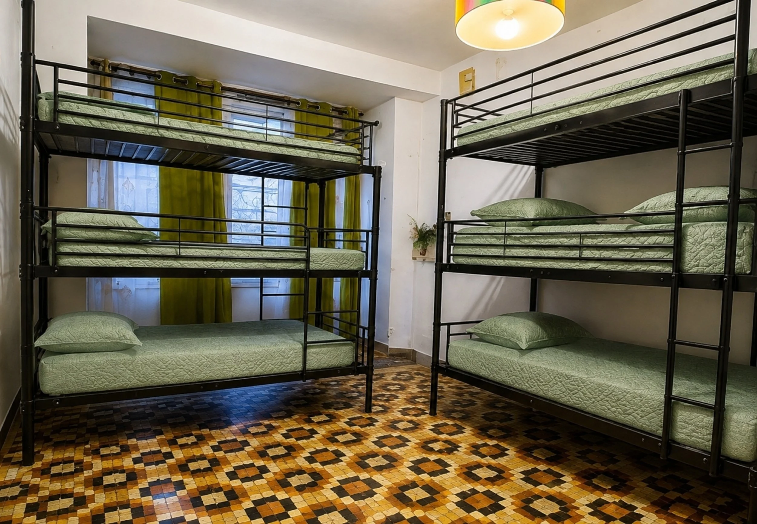 Chambres avec lits superposés en métal noir, matelas et oreillers verts, rideaux verts et volets bleus derrière la fenêtre, sol en carreaux de mosaïque, lampe suspendue au plafond.