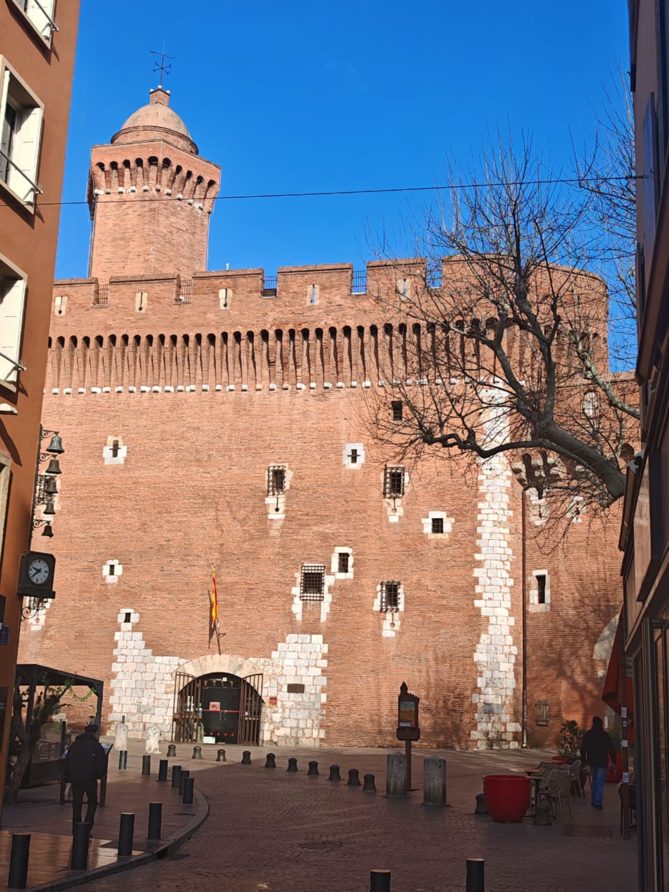 Une vieille forteresse en briques rouges avec une tour, des petites fenêtres, et une porte d'entrée, située dans une rue pavée avec quelques piétons.
