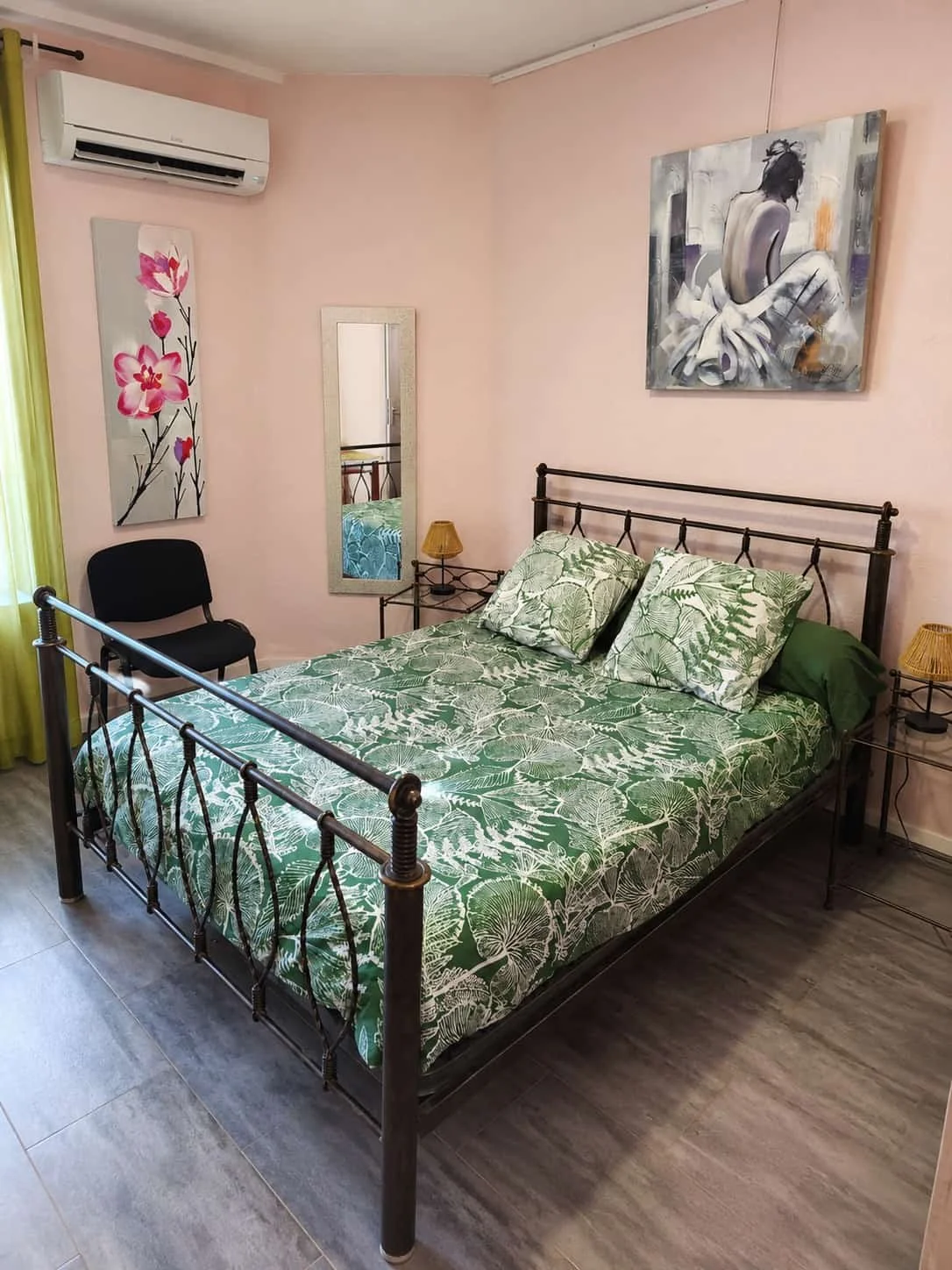 Chambre avec lit double à cadre en métal noir, literie avec motif de feuilles vertes, deux oreillers assortis, deux lampes de chevet en rotin avec abat-jour jaune, tableau abstrait de femme assise en arrière-plan, peinture florale sur le mur, miroir rectangulaire, chaise noire, climatiseur mural, rideaux verts sur la fenêtre, sol en carrelage gris.