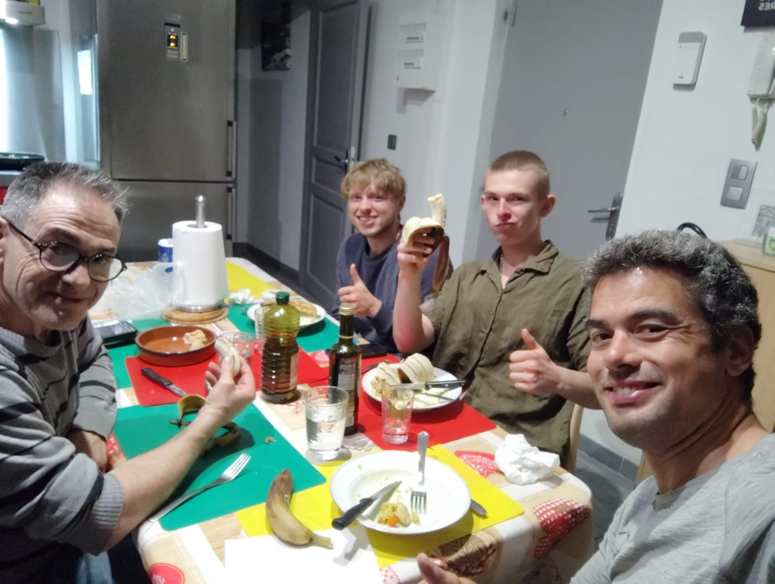 Quatre personnes autour d'une table à manger, souriantes, avec des morceaux de banane, une bouteille d'huile et un film de cuisine en arrière-plan.