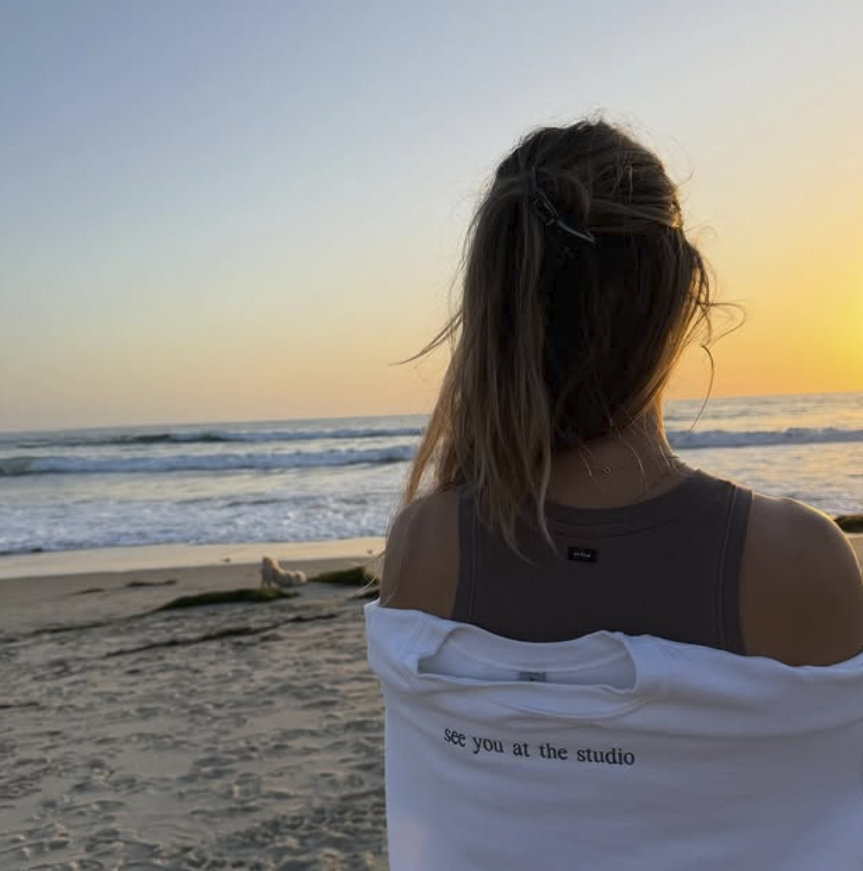 An image of a woman at the beach wearing The Pilates Studio merch that says "see you at the studio"
