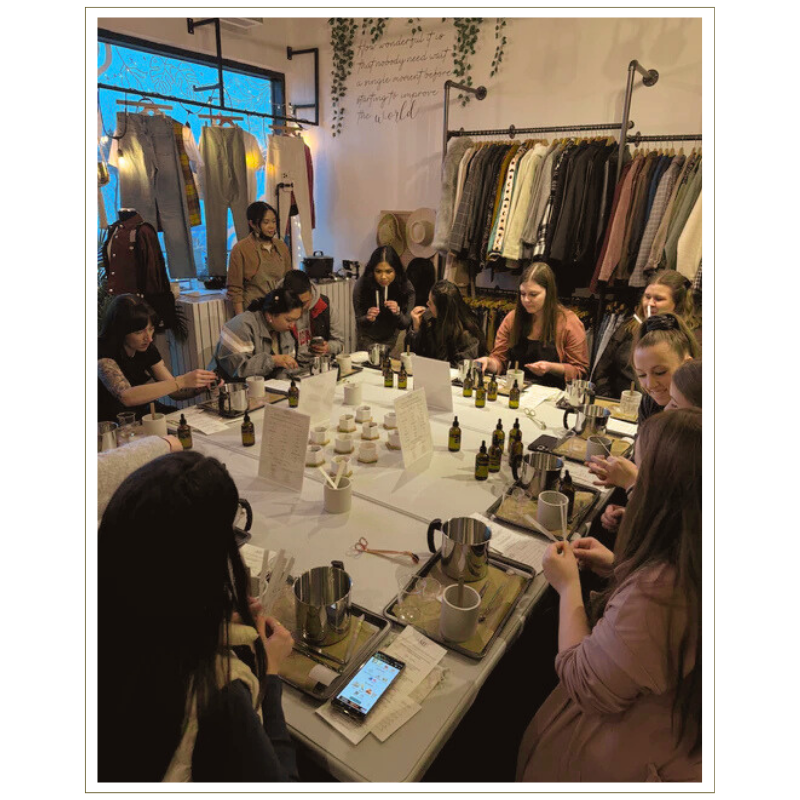 Group of women participating in a candle-making workshop around a large table with candles, supplies, and scent bottles inside a clothing boutique.