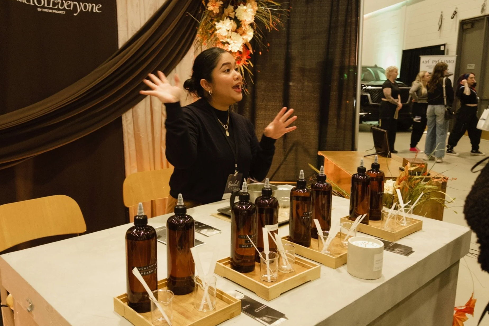 A woman with dark hair pulled back, gesturing with her hands, standing behind a table with amber fragrance oil bottles and fragrance strips at an event.