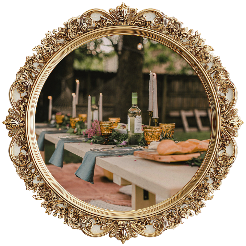 An outdoor dinner table set with a light blue tablecloth, candelabras with lit candles, wine bottles, glasses, and a plate of bread, with trees and a wooden fence in the background.
