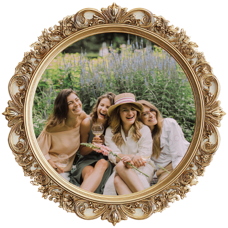 Four women sitting outdoors in front of a purple flowering plant, smiling and laughing, holding wine glasses, with one wearing a hat.