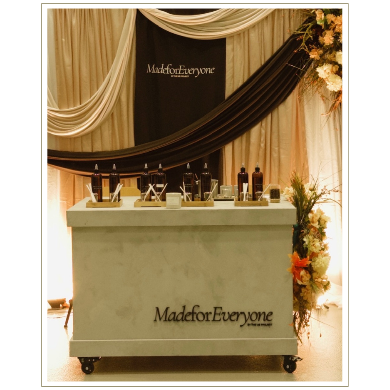 A diffuser and candle-making cart with several amber bottles of fragrance oils set against a decorated backdrop with beige and dark fabric drapes and floral arrangements. The cart has the words 'Made for Everyone' on it.