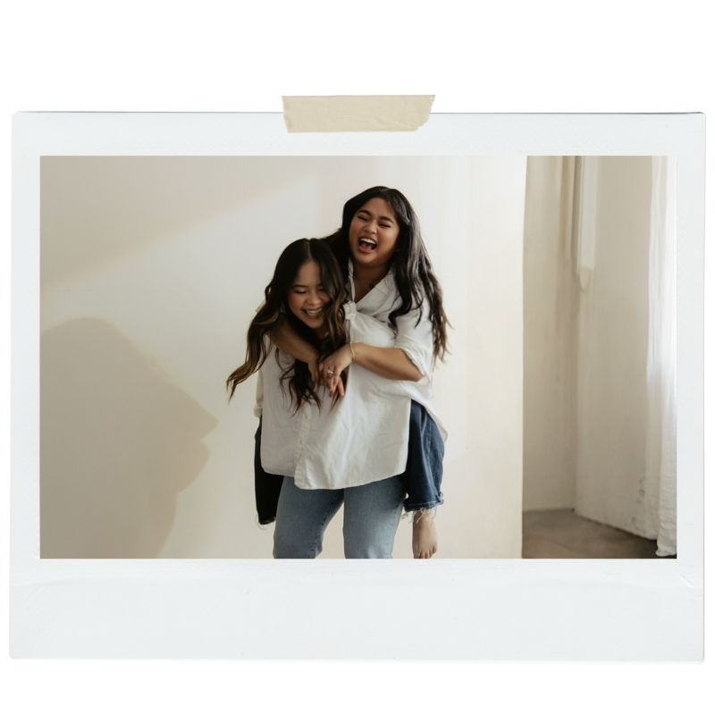 Two women laughing and hugging each other playfully in a bright room, one carrying the other on her back.