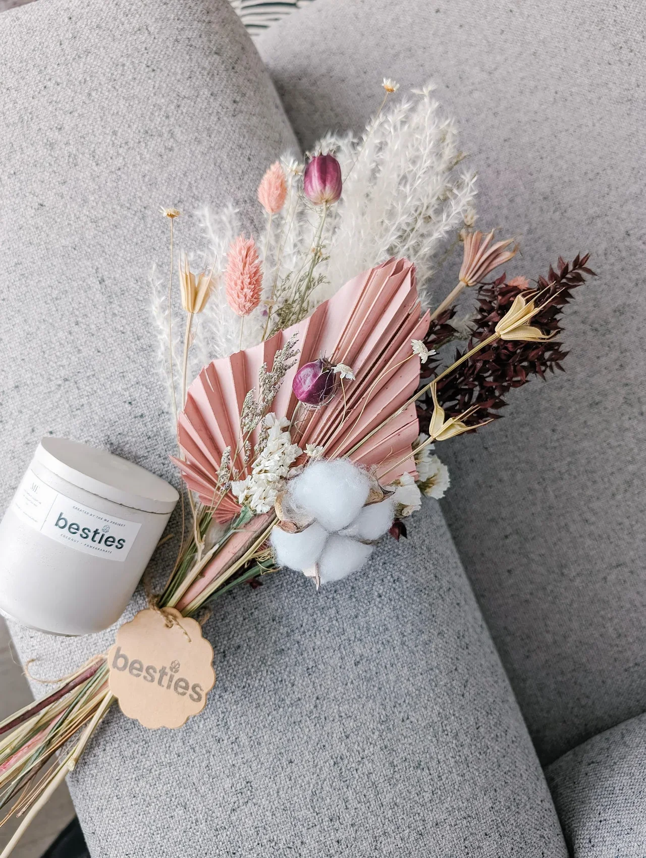A bouquet of dried flowers resting on a gray sofa, along with a gray concrete candle labeled 'besties' and a small round tag with 'besties' written on it.