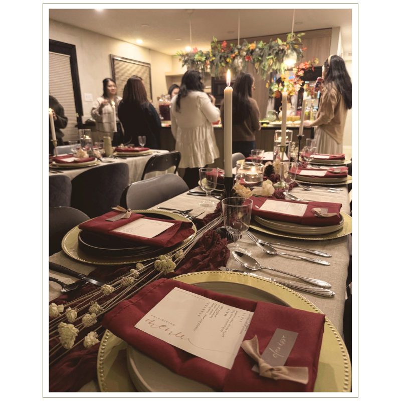 A dinner table set with gold-rimmed plates, red cloth napkins, glassware, and silverware, decorated with a centerpiece of flowers and candles, with people standing in the background.