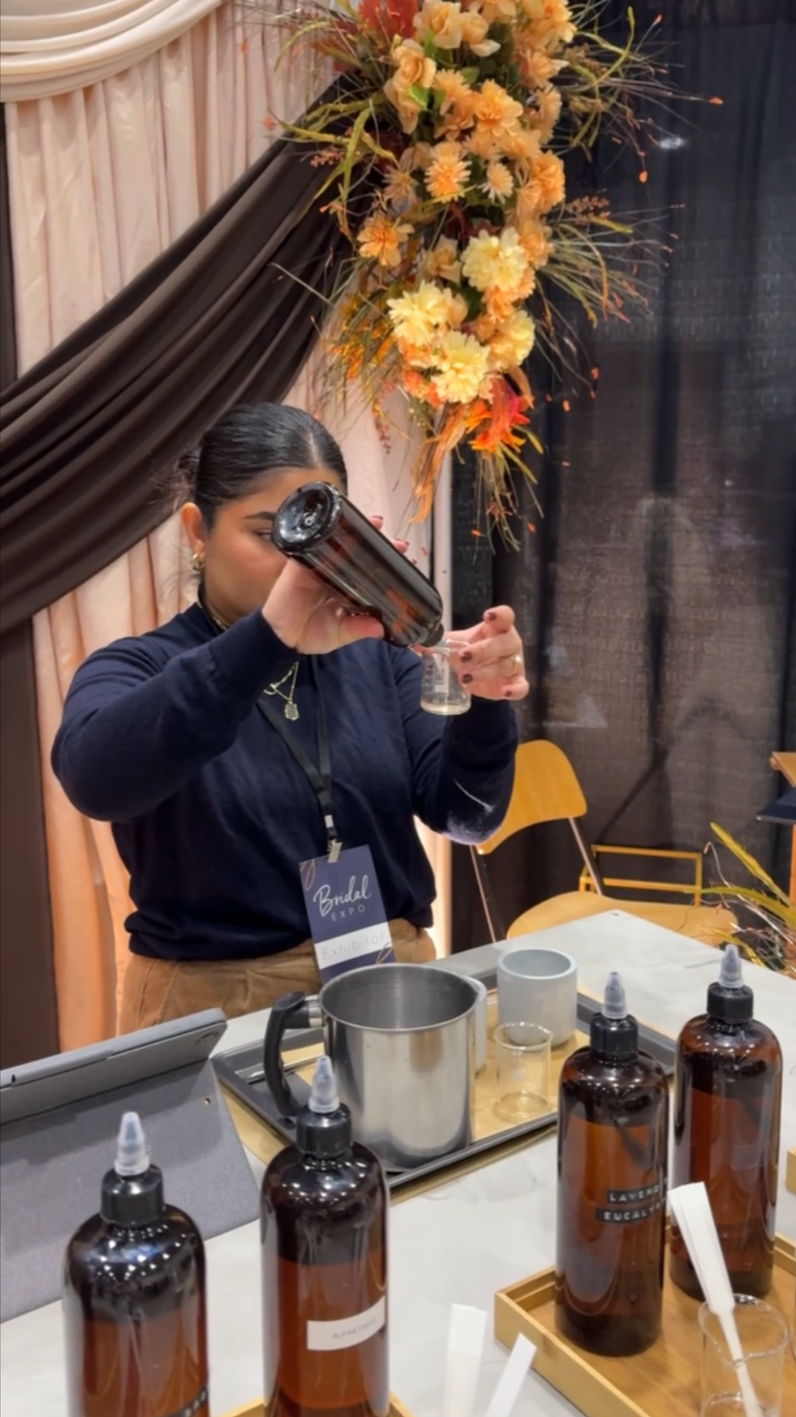 A woman at a booth pouring fragrance oil from an amber bottle into a beaker at an event with fall vibe decor, including large flower arrangements with orange, yellow, and brown foliage.