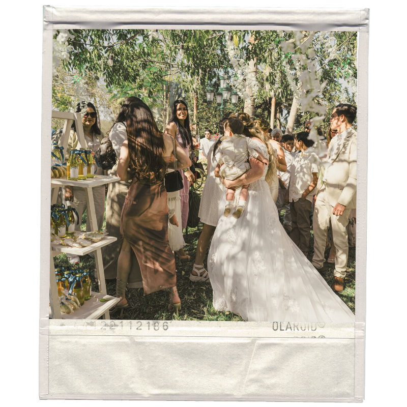 People gathered outdoors at a celebration, including a woman in a wedding dress holding a child, surrounded by friends and family on a sunny day with trees and greenery in the background.