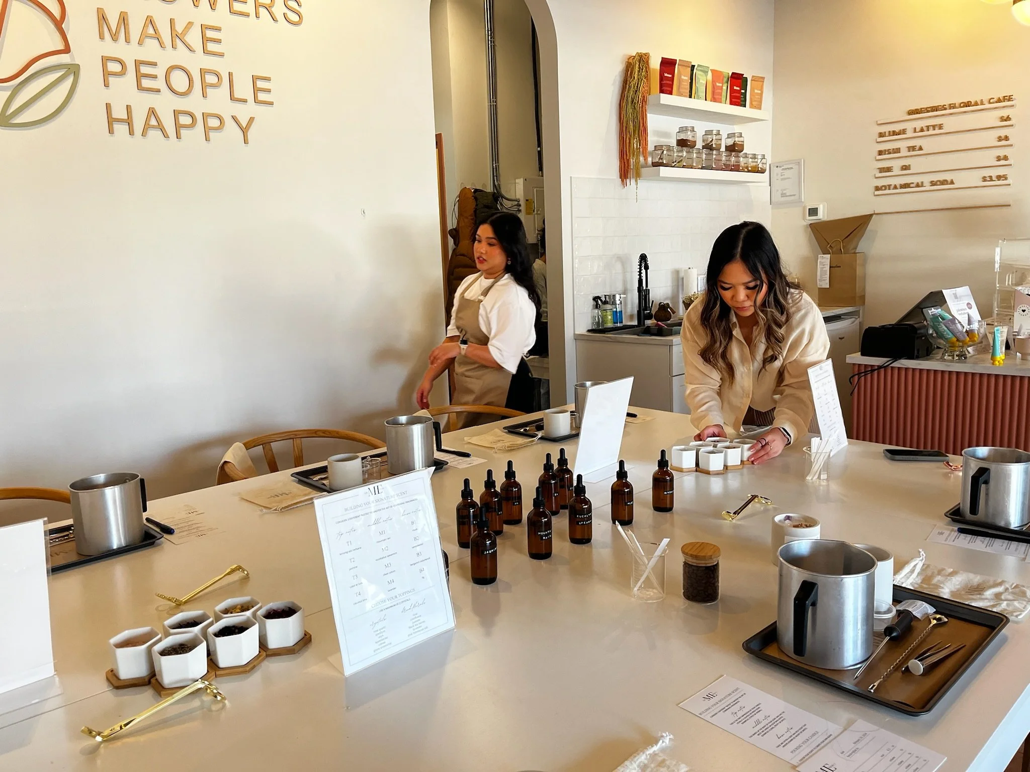 Inside a floral café, two women with dark hair and light clothing setting up for a candle-making workshop, including fragrance oil bottles, trays, and other candle-making tools.