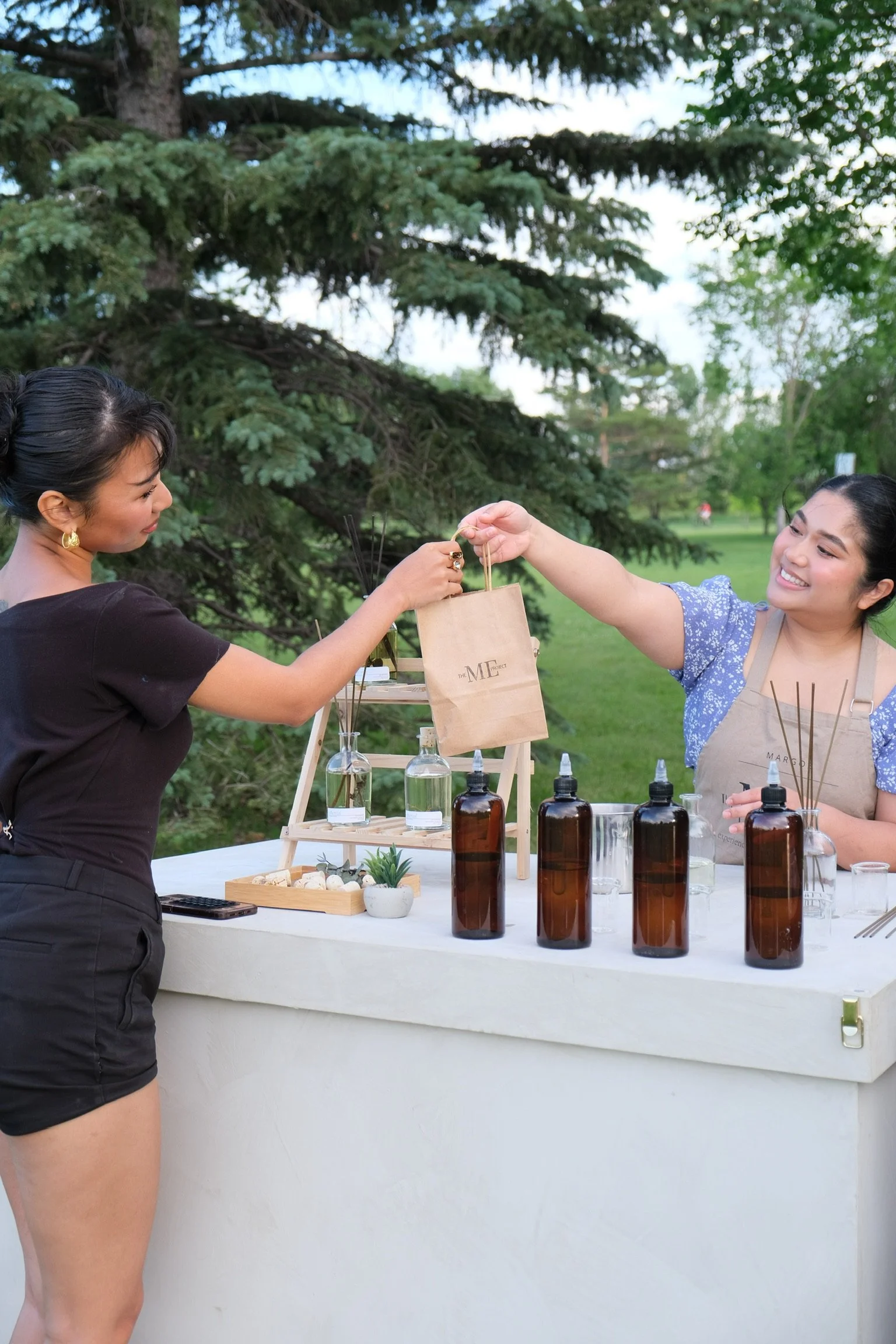 Two women at an outdoor reed diffuser cart exchanging a paper bag, with bottles of fragrance oils and reed diffusers on the cart, in a park-like setting with trees and grass in the background.