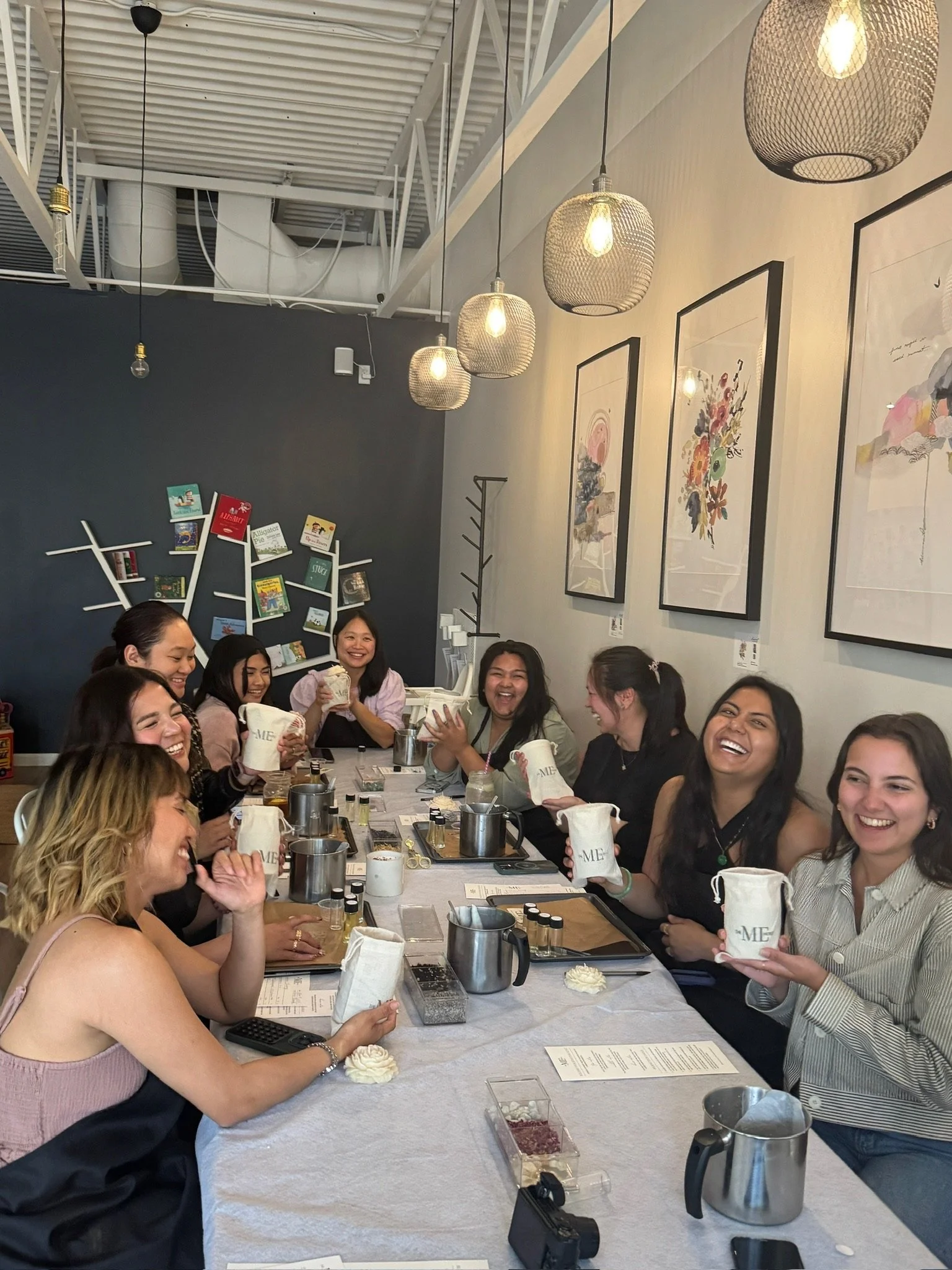 Group of women sitting around a table at a coffee shop, smiling, holding their customized candles they just made, with candle-making trays and tools on the table.
