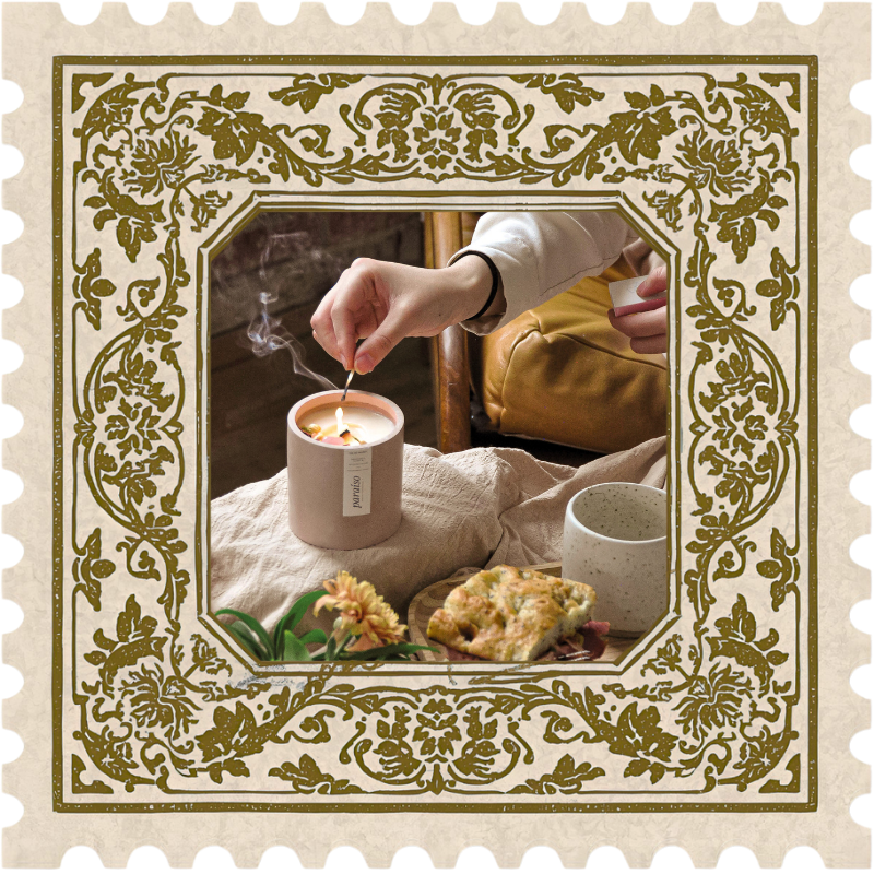 A photo of a person lighting a candle on a cozy table with a slice of cake, a mug, and a flower within an stamp-shaped frame with ornate borders.
