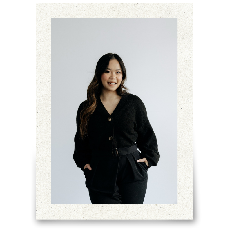 A young woman with long, wavy dark hair wearing a black cardigan and black trousers, smiling against a plain white background.