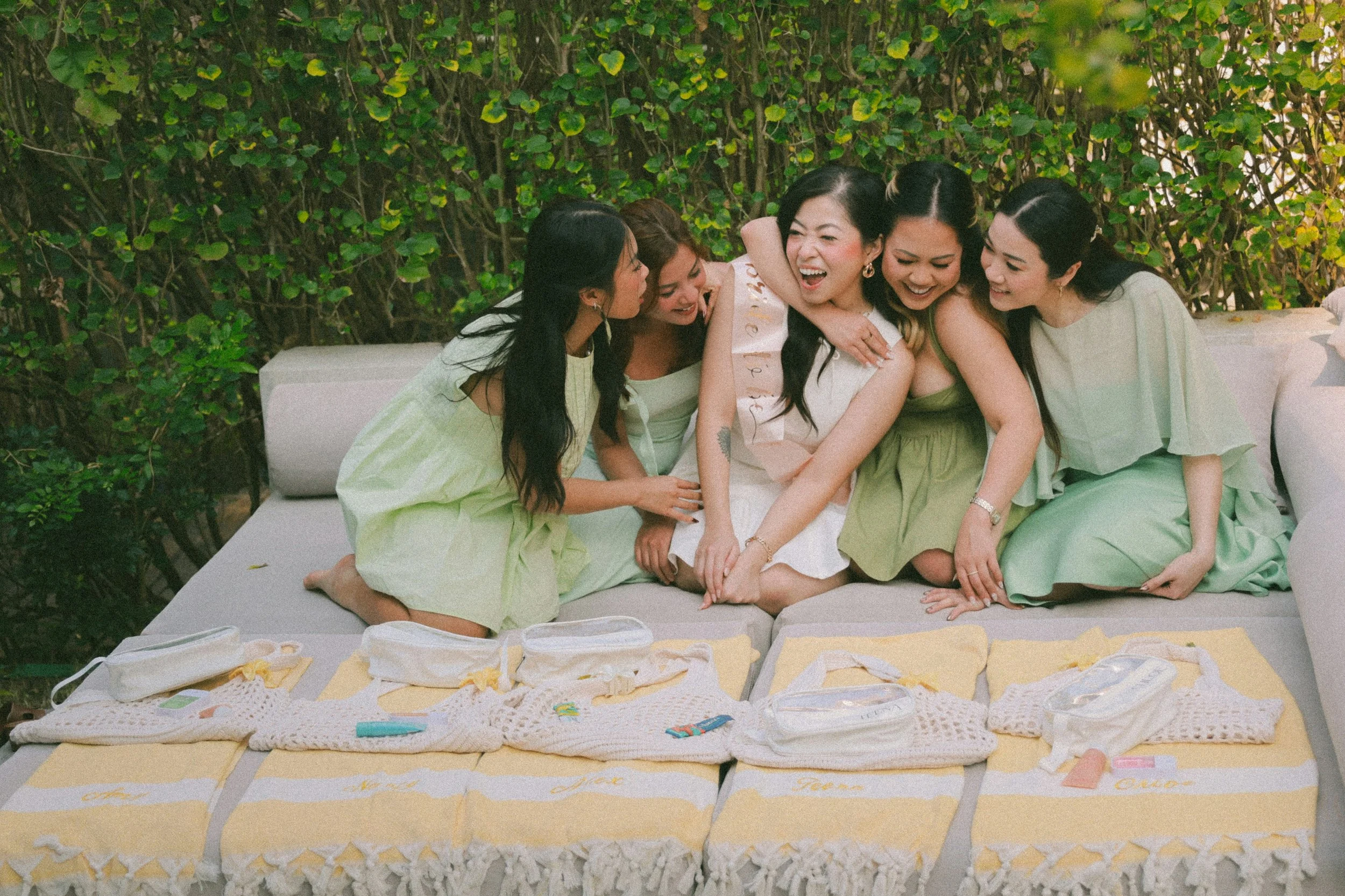 Six women sitting on a cushioned outdoor bench, smiling and laughing together during a celebration. They are dressed in pastel green and white dresses. In front of them are yellow towels with wedding favours.