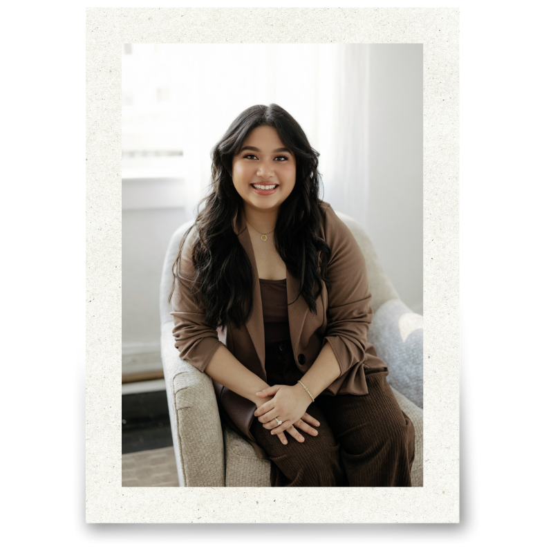 Young woman with long dark hair, smiling, sitting on light-colored armchair in a bright room with white curtains.