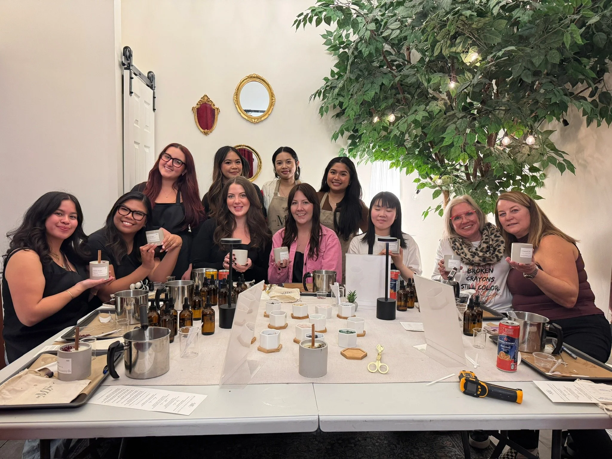 Group of women gathered around a table participating in a candle-making workshop, with various candles, tools, and supplies on the table, and a large plant and wall mirrors in the background.