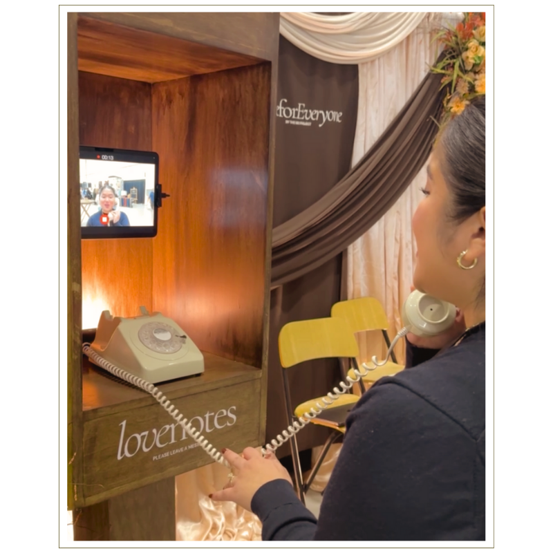 A woman holding a vintage rotary telephone receiver and speaking into a retro-style corded phone mounted on a wooden stand with a tablet showing a video call. The stand has a sign that reads "lovenotes" and is decorated with flowers and draped fabric in the background.