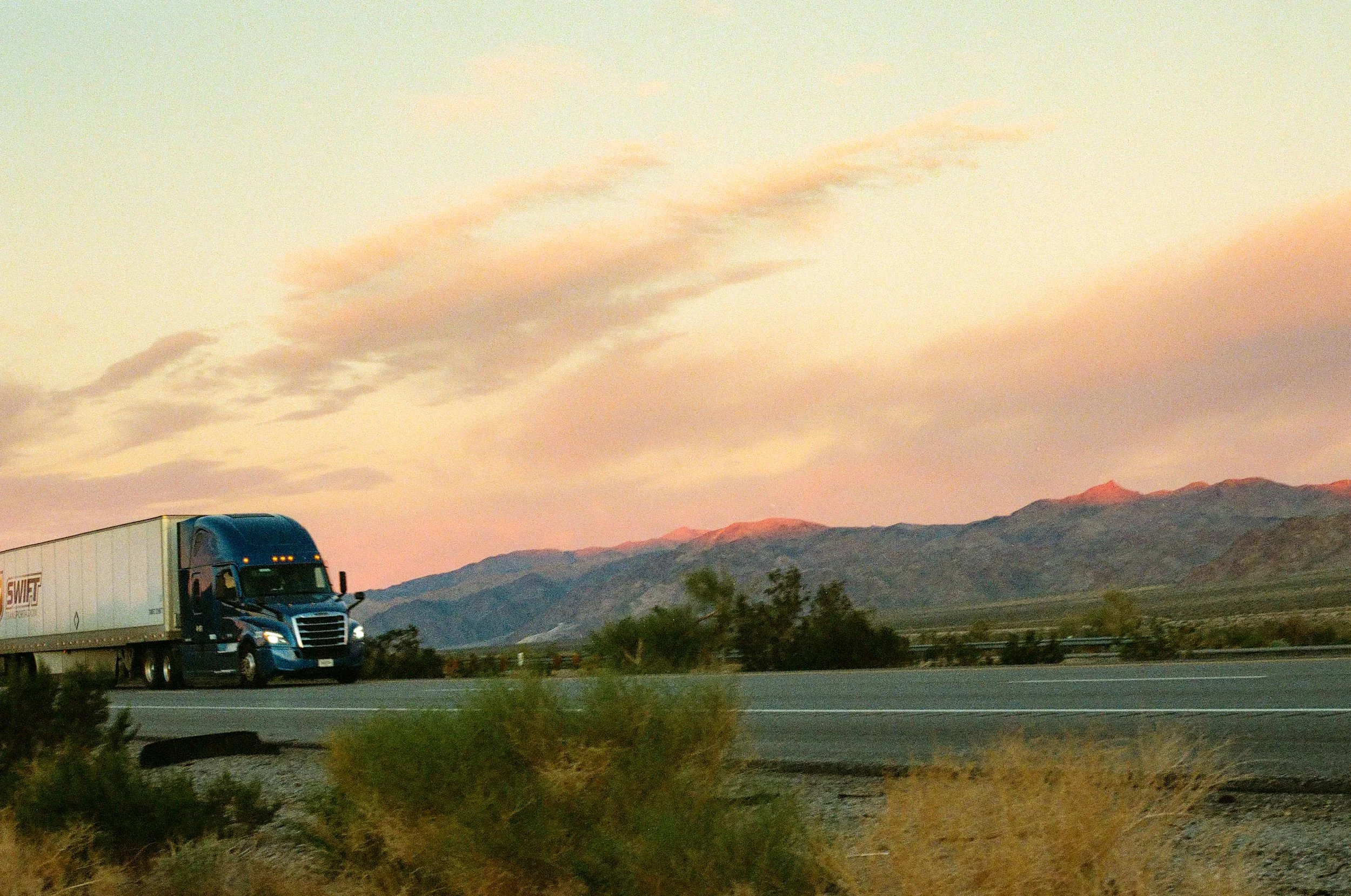 A semi-truck driving along a highway with a mountainous landscape and pinkish sky in the background at sunset.