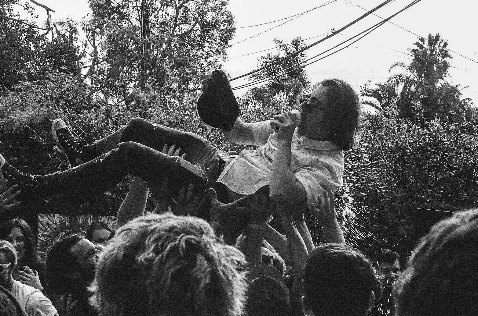 A woman in sunglasses and a white shirt is being lifted by a crowd, sipping from a bottle, with trees and power lines in the background.