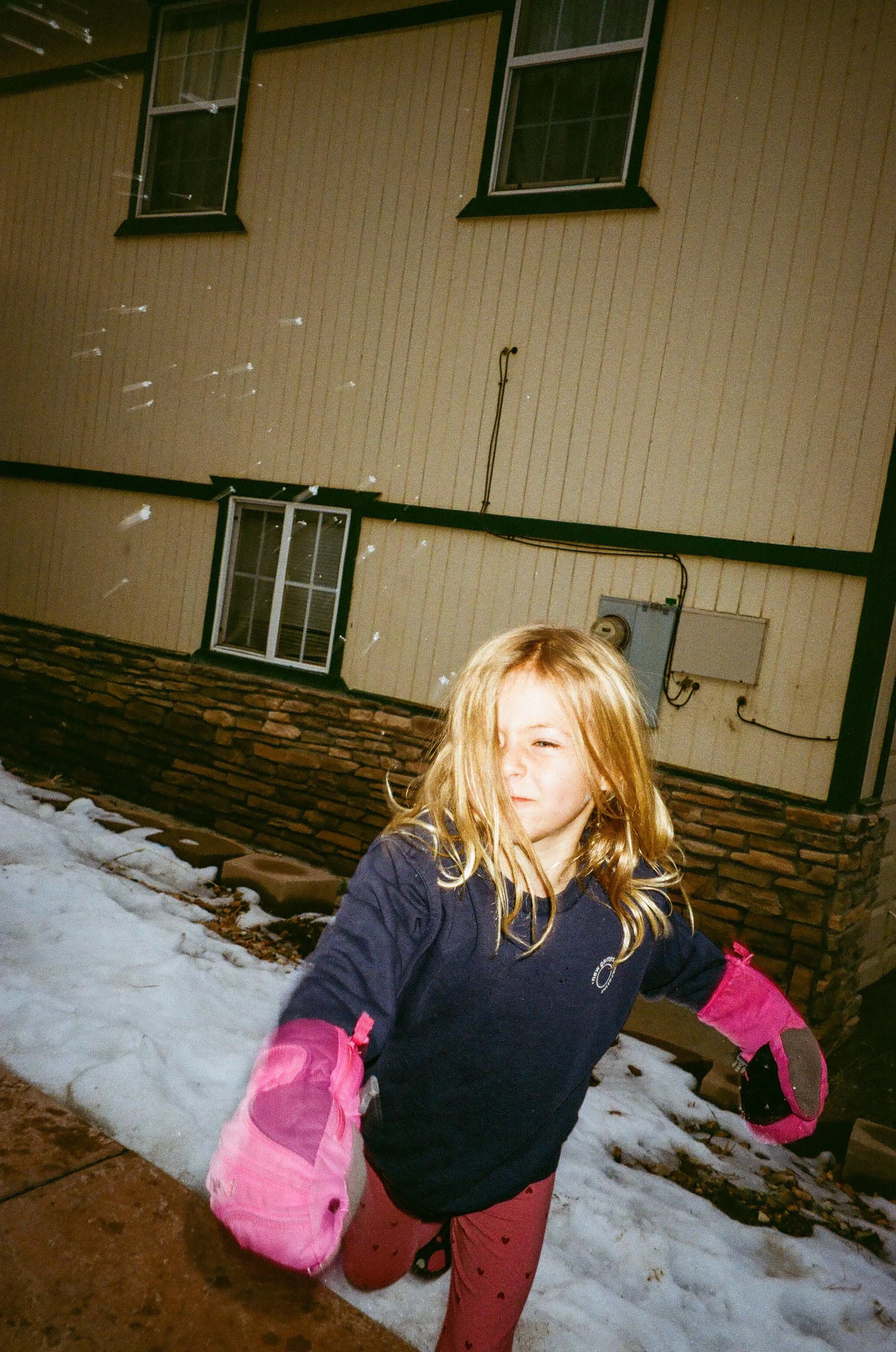 A young girl with blonde hair running outdoors near a house with beige siding and brick foundation. She is wearing pink pants, a navy blue jacket, and pink gloves, with snow on the ground.