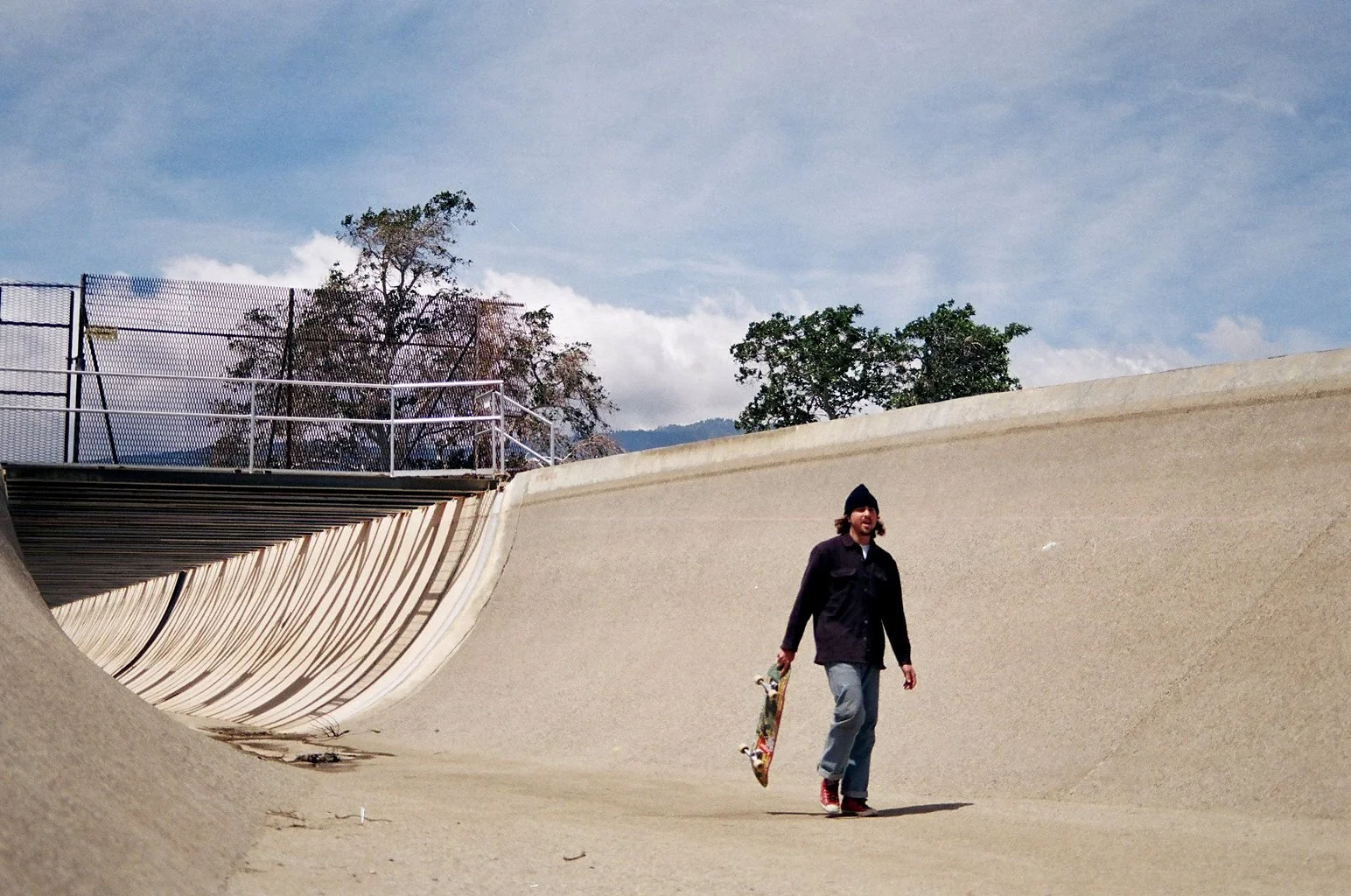 A man in a black jacket, gray pants, and a black beanie is walking with a skateboard in a concrete skatepark under a partly cloudy sky, with trees in the background.