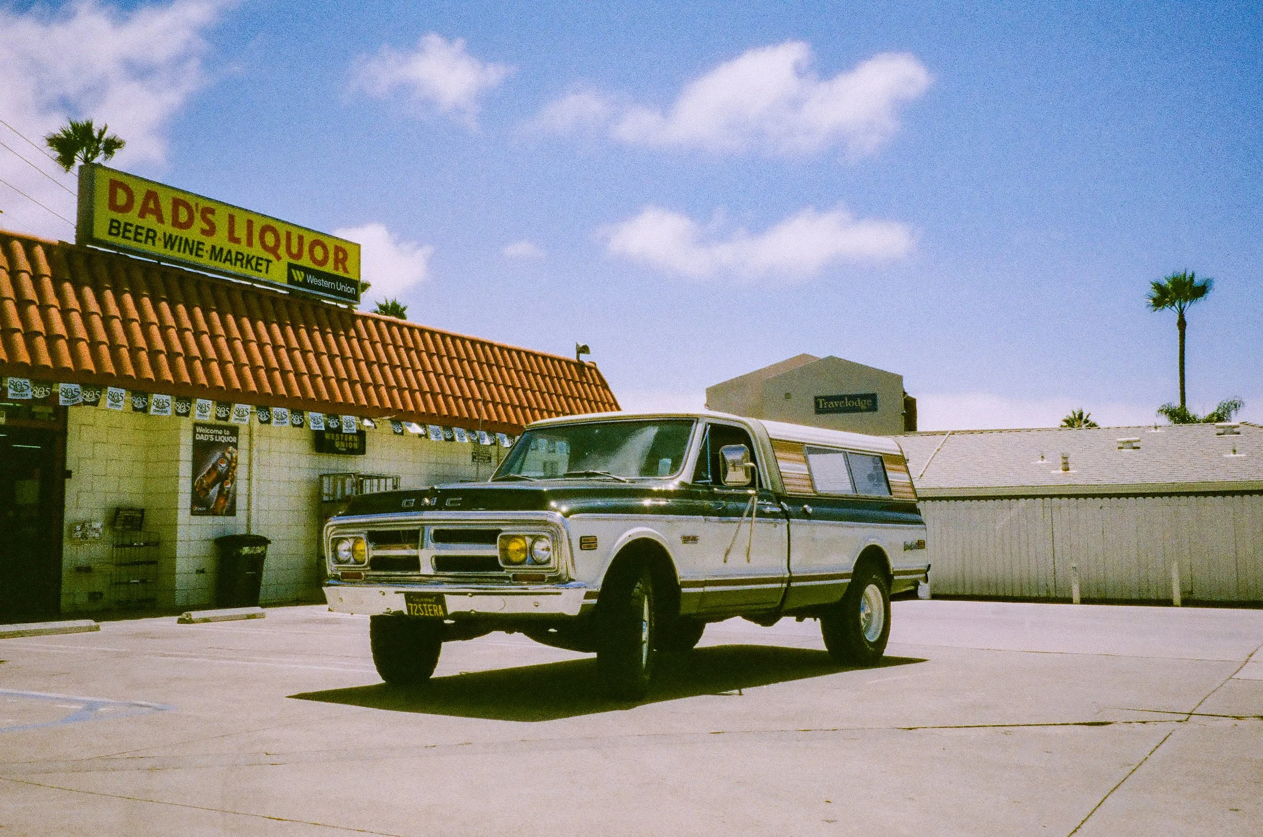 A vintage GMC pickup truck parked in front of a liquor store named Dad's Liquor with a red-tiled roof, under a blue sky with a few clouds, and palm trees in the background.