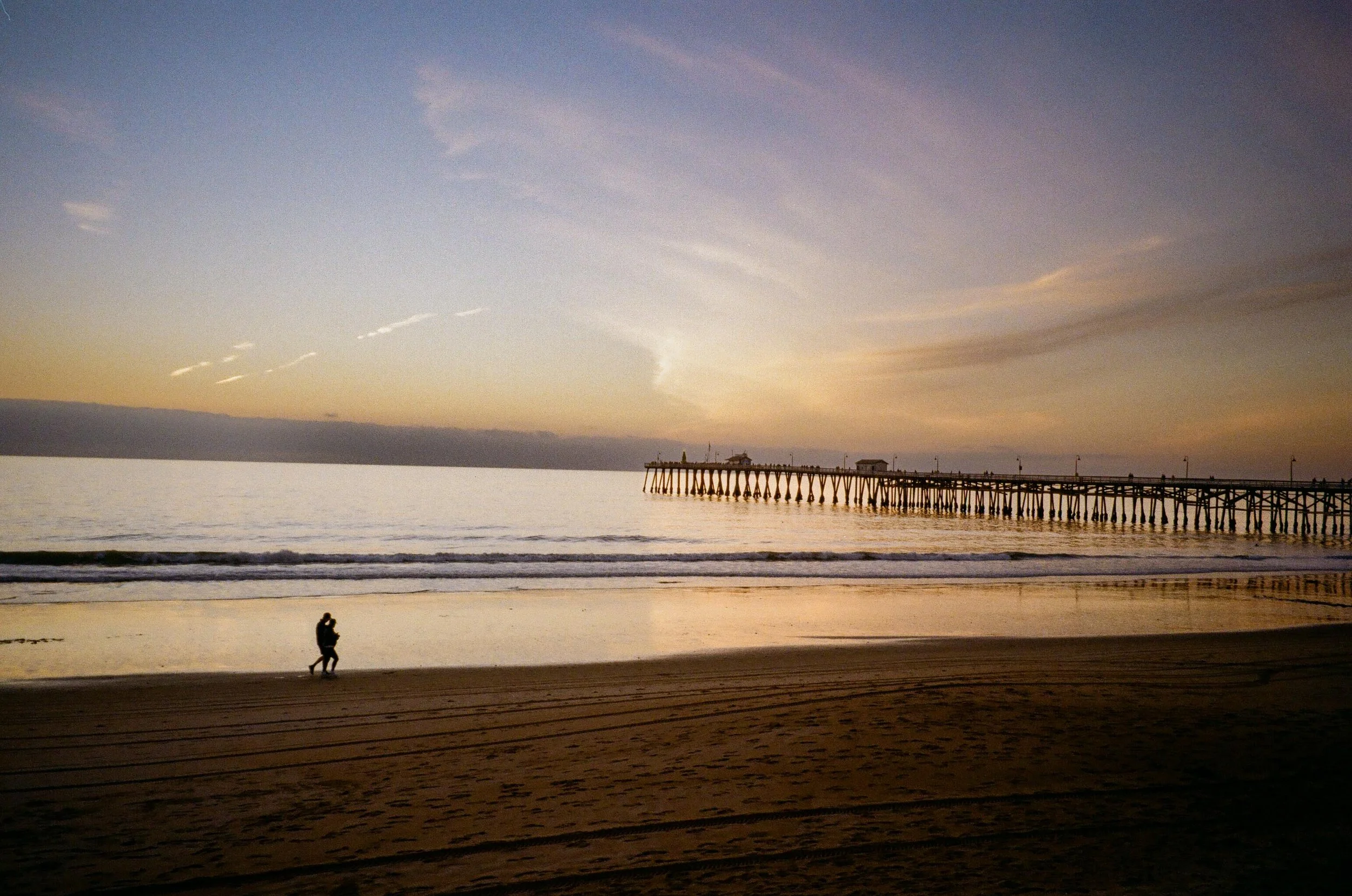 A sunset at the beach with a person walking along the shore and a pier extending into the water.