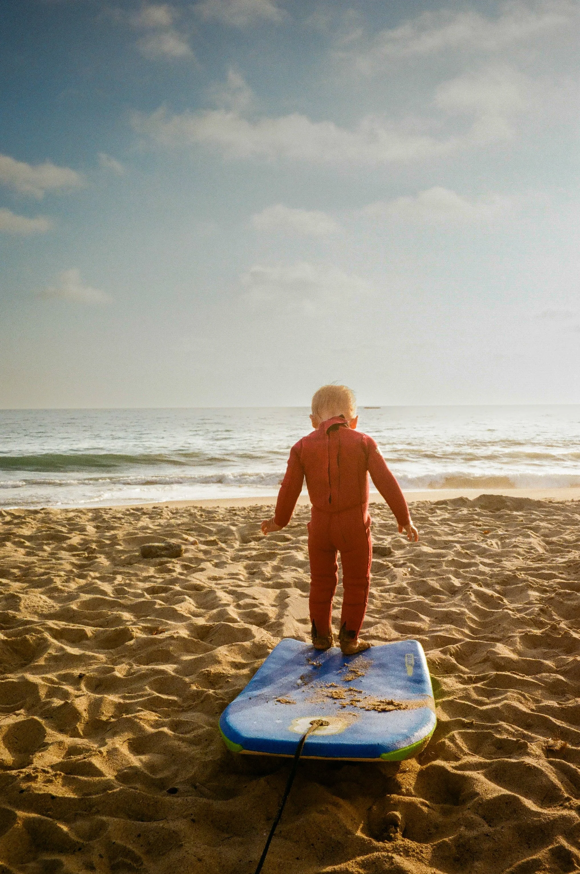 Child standing on a blue surfboard on the sandy beach, facing the ocean with a cloudy sky overhead.
