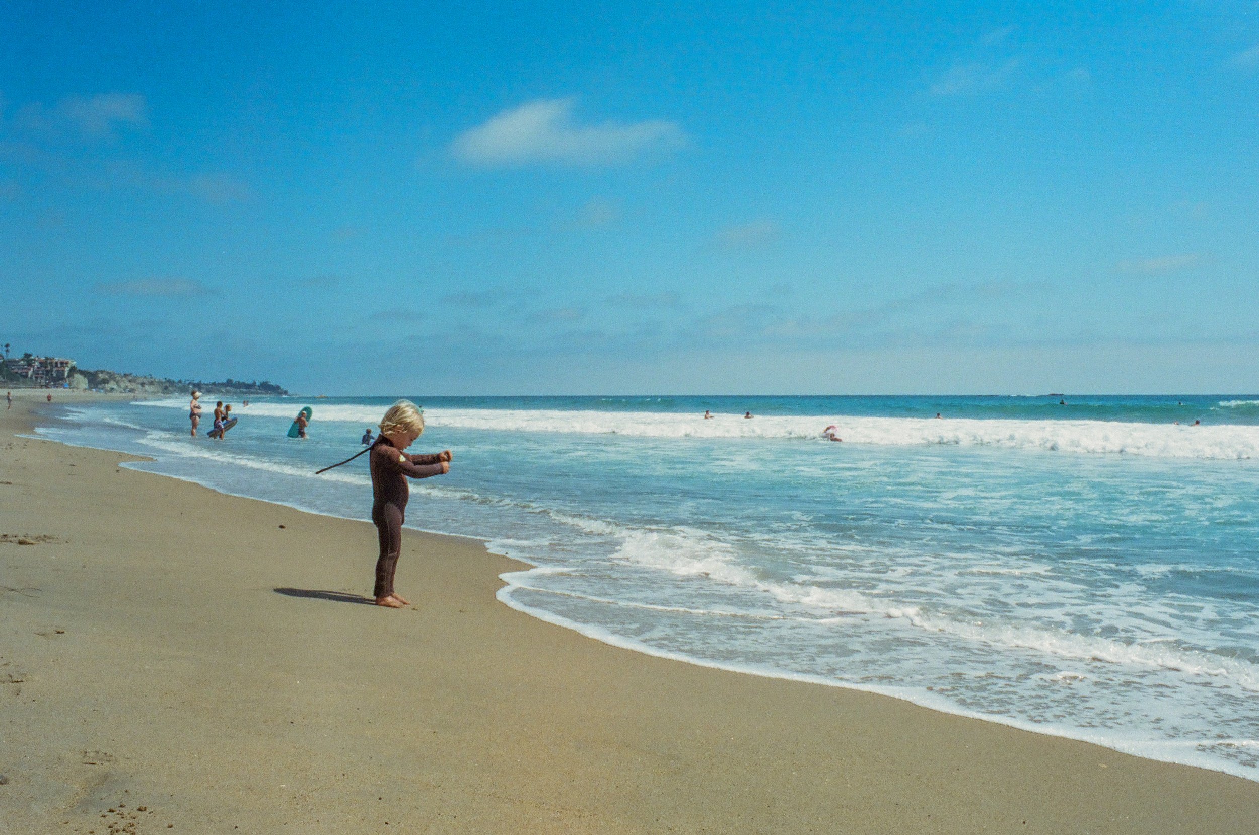 A young child in a brown wetsuit stands on the sandy beach near the water, with people swimming and playing in the ocean under a blue sky.