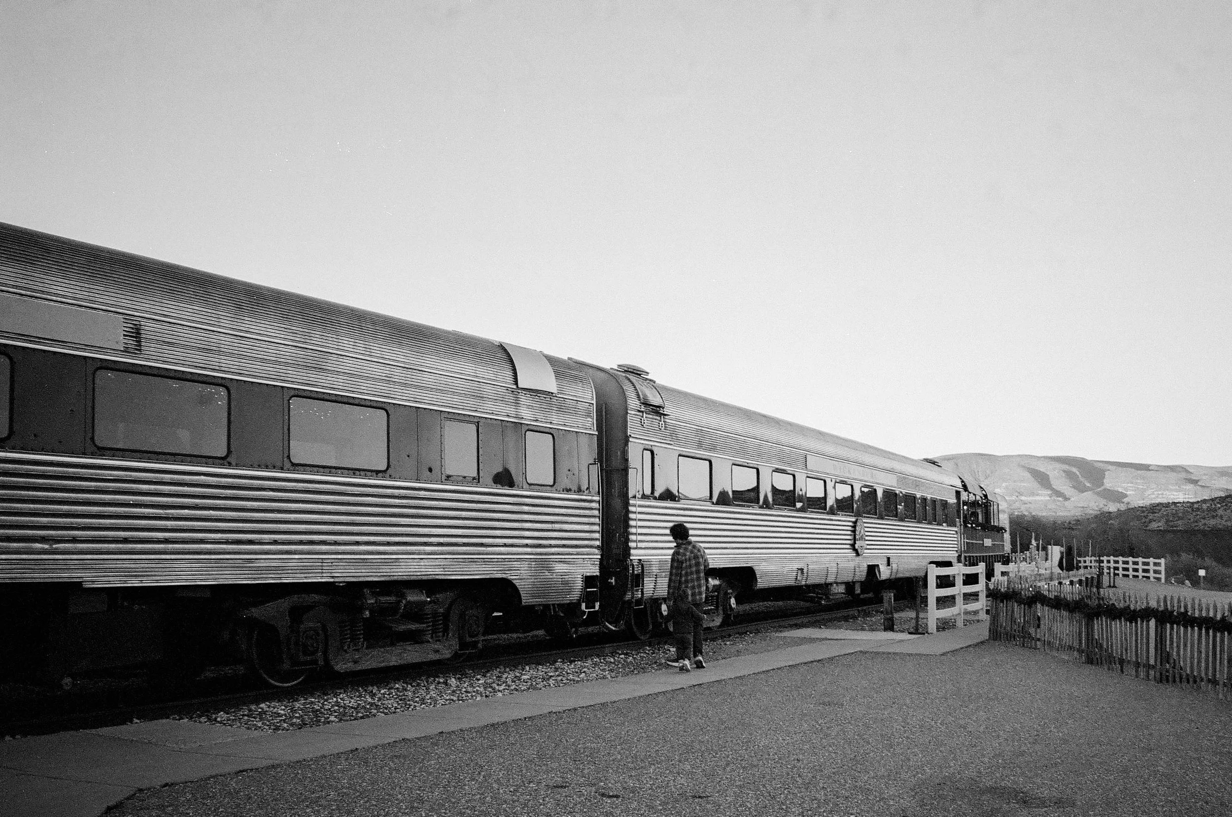 A person standing near a vintage train on train tracks in a rural area, with hills in the background.