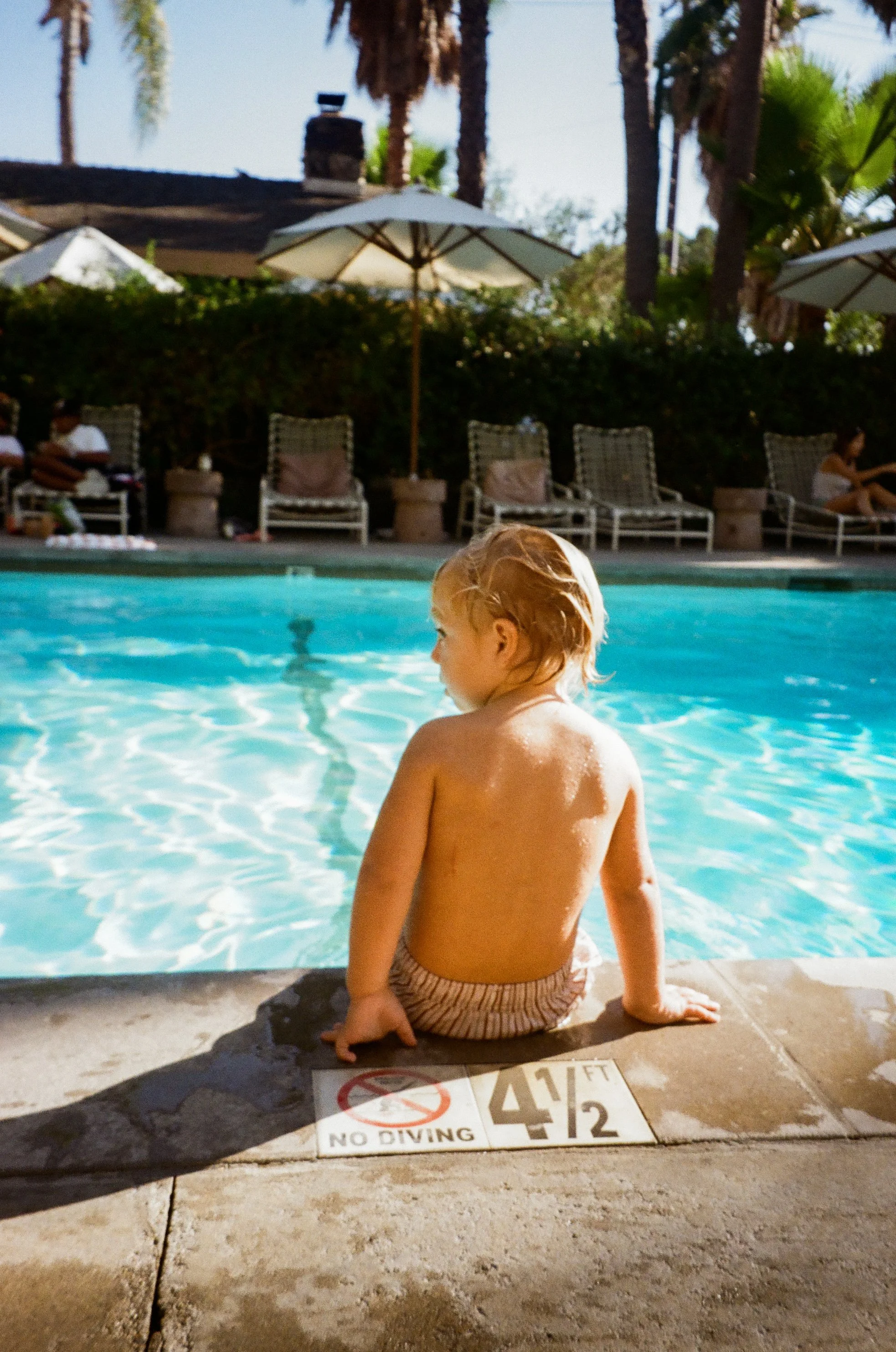 A young child with blonde hair sitting on the edge of a swimming pool, wearing striped swim trunks, with a 'No Diving' sign on the pool deck nearby.