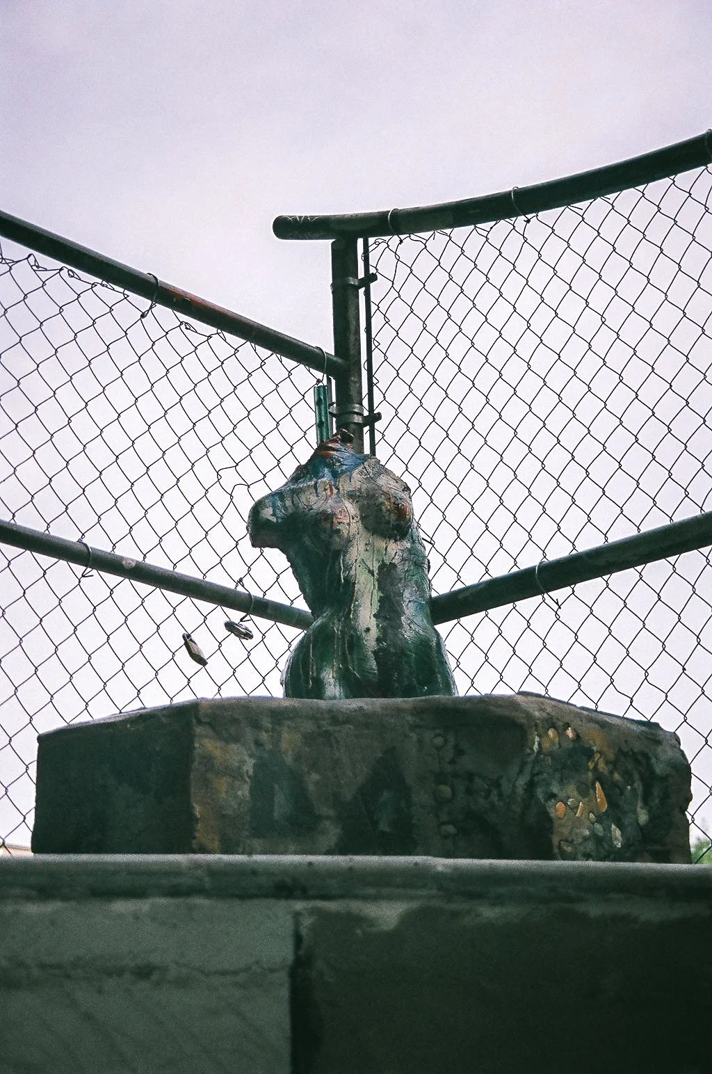 A close-up of a small sculpture of a dog on a stone pedestal, behind a chain-link fence on a cloudy sky day.