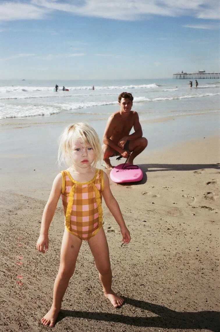 A young blonde girl in a pink and yellow checkered swimsuit standing on the sand at the beach, with a shirtless young man kneeling in the background near the water with a pink boogie board, and the ocean with a pier extending into the water in the distance.