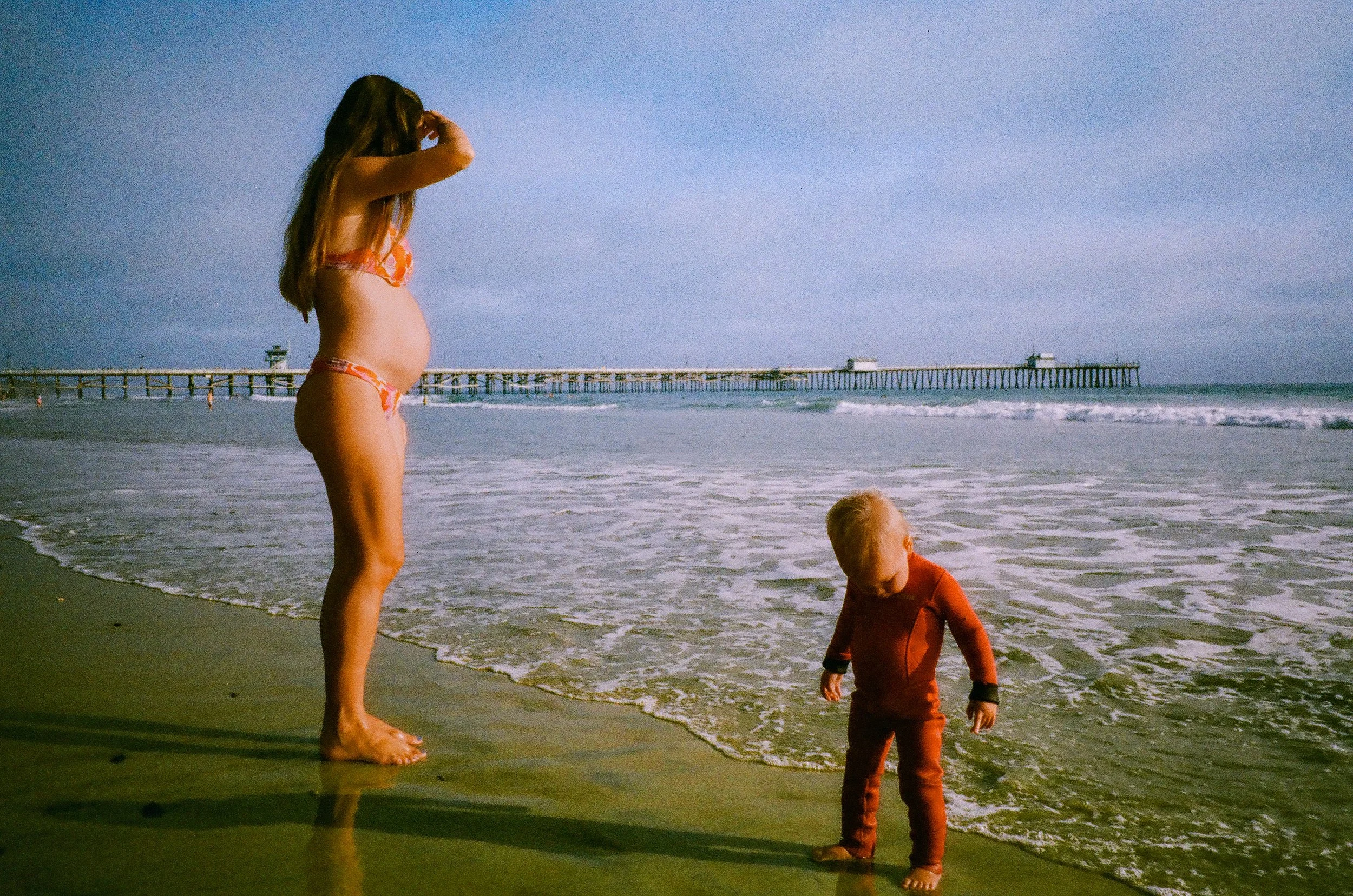 A woman in a bikini and a young child in a wetsuit standing on a beach near the water with a pier extending into the ocean in the background.