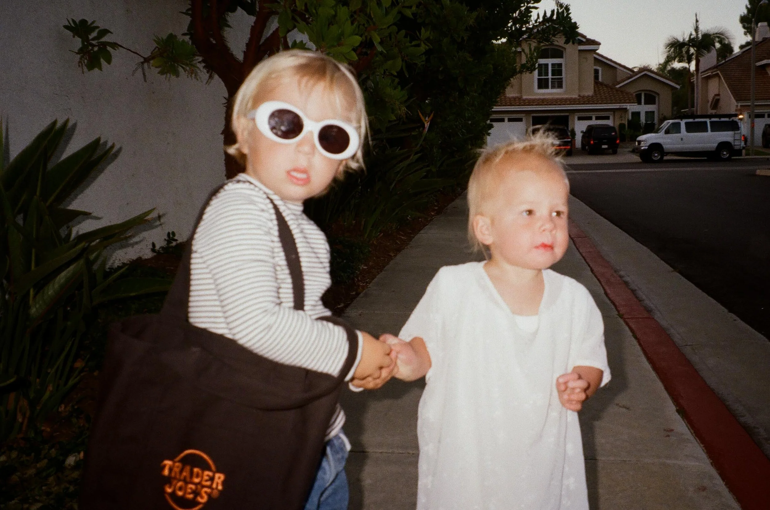 Two young children walking outside on a sidewalk, holding hands, with houses and parked cars in the background. The older child wears white sunglasses and a black and white striped shirt, carrying a Trader Joe's grocery bag. The younger child wears a white dress.