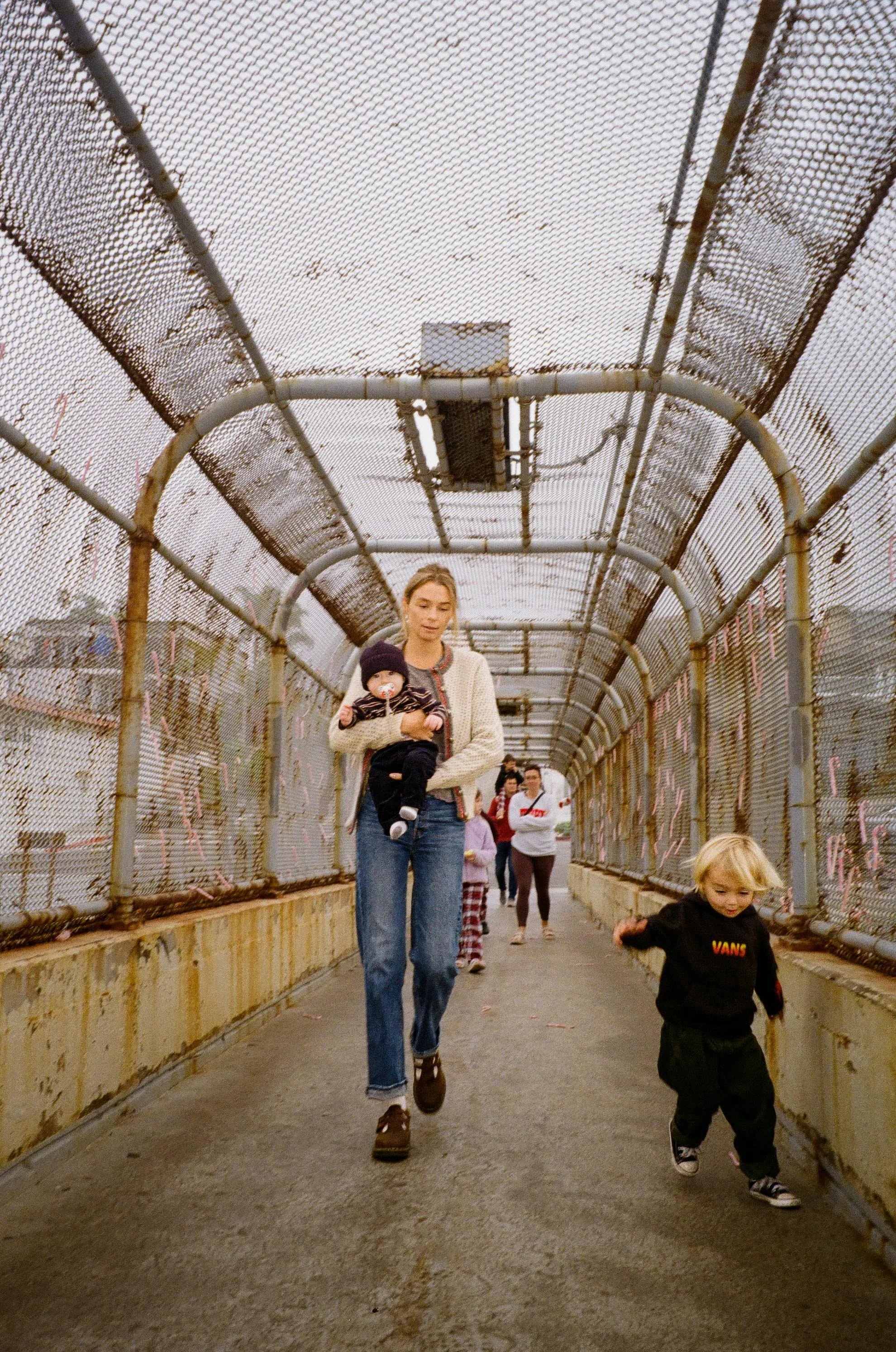 People walking on a fenced overpass, including a woman holding a child and a young boy running.