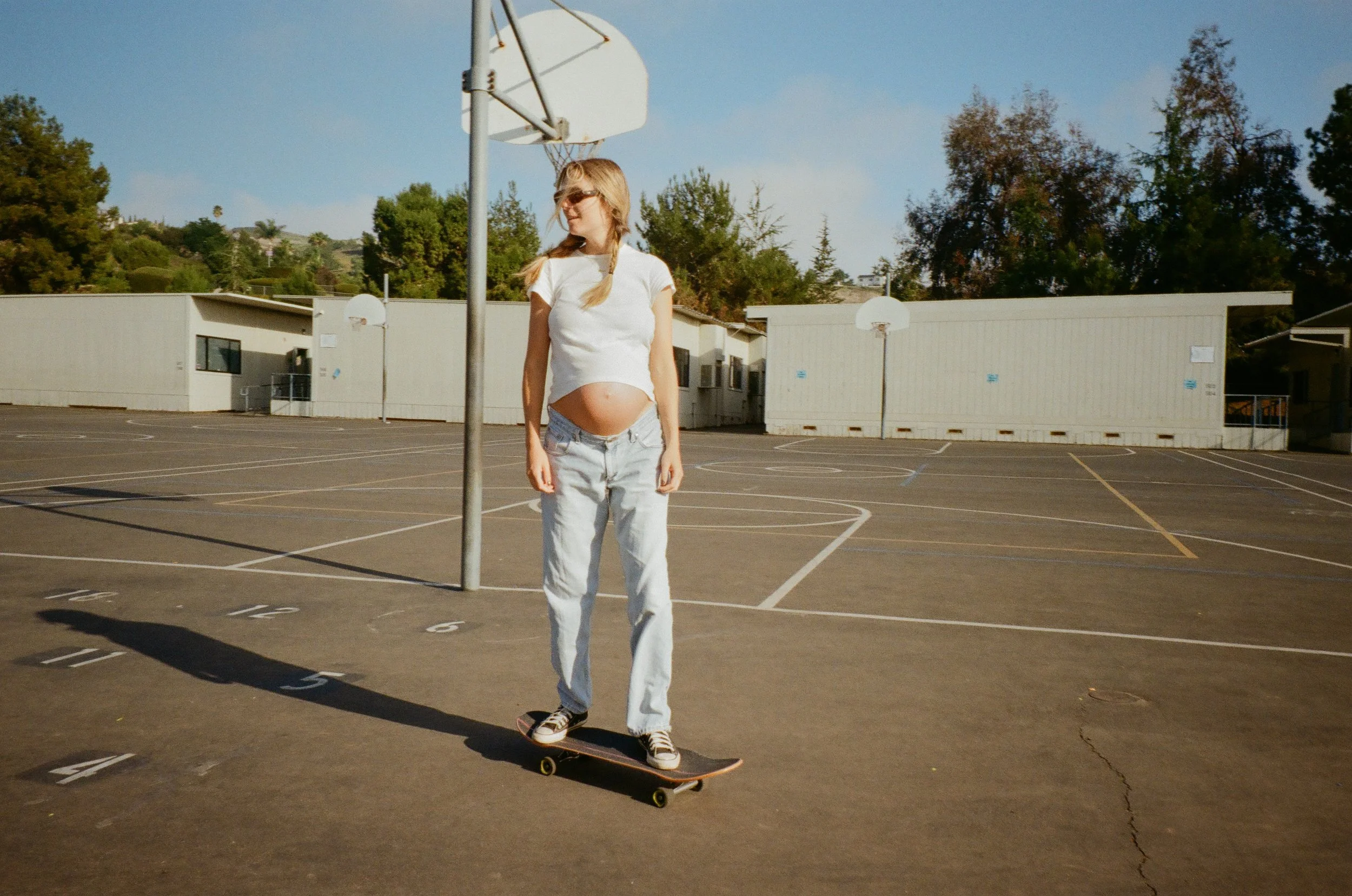 A pregnant woman skateboarding on an outdoor basketball court.
