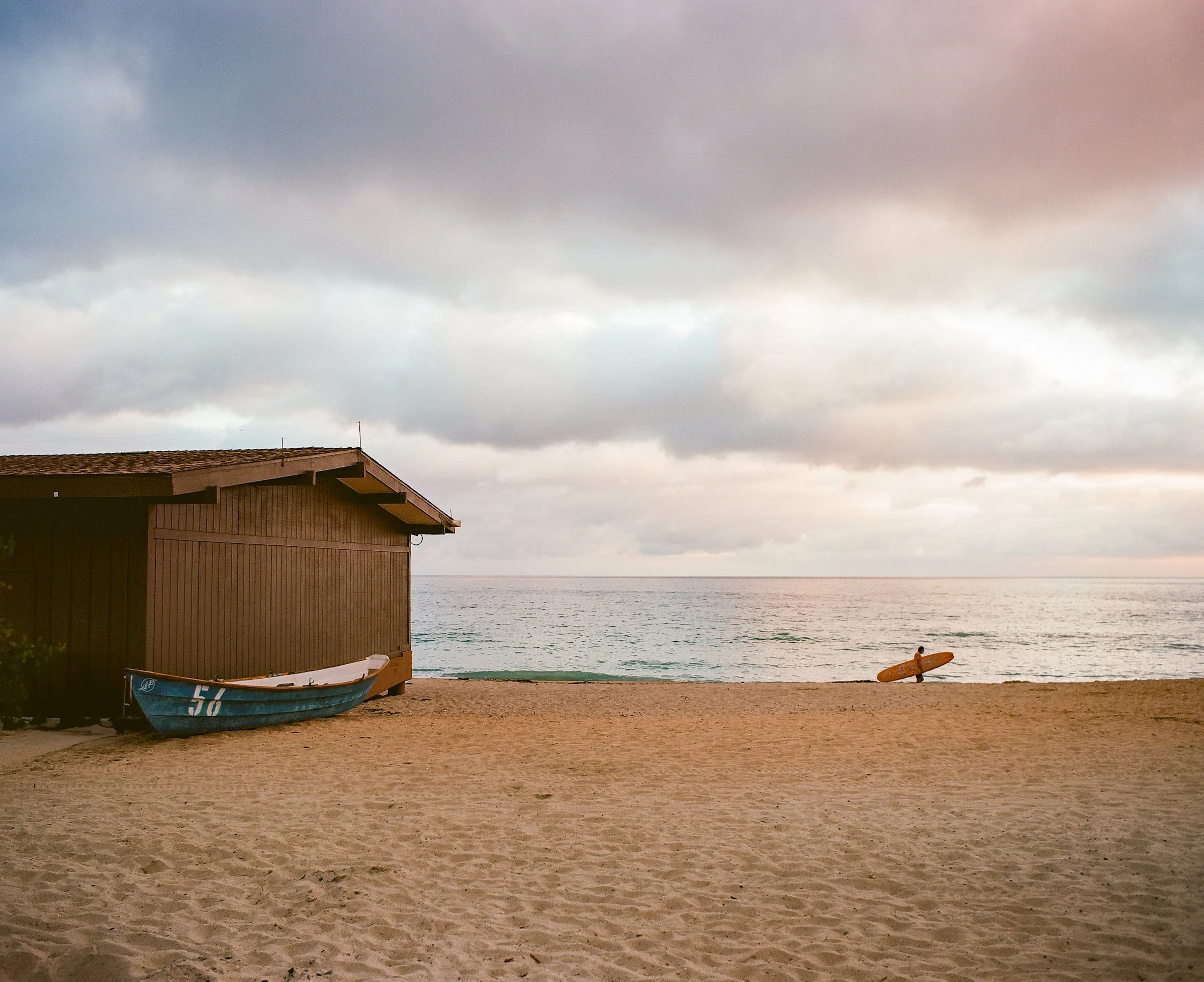 A sandy beach with a brown wooden beach house, a small blue boat, and a person with a surfboard near the water, under a cloudy sky.