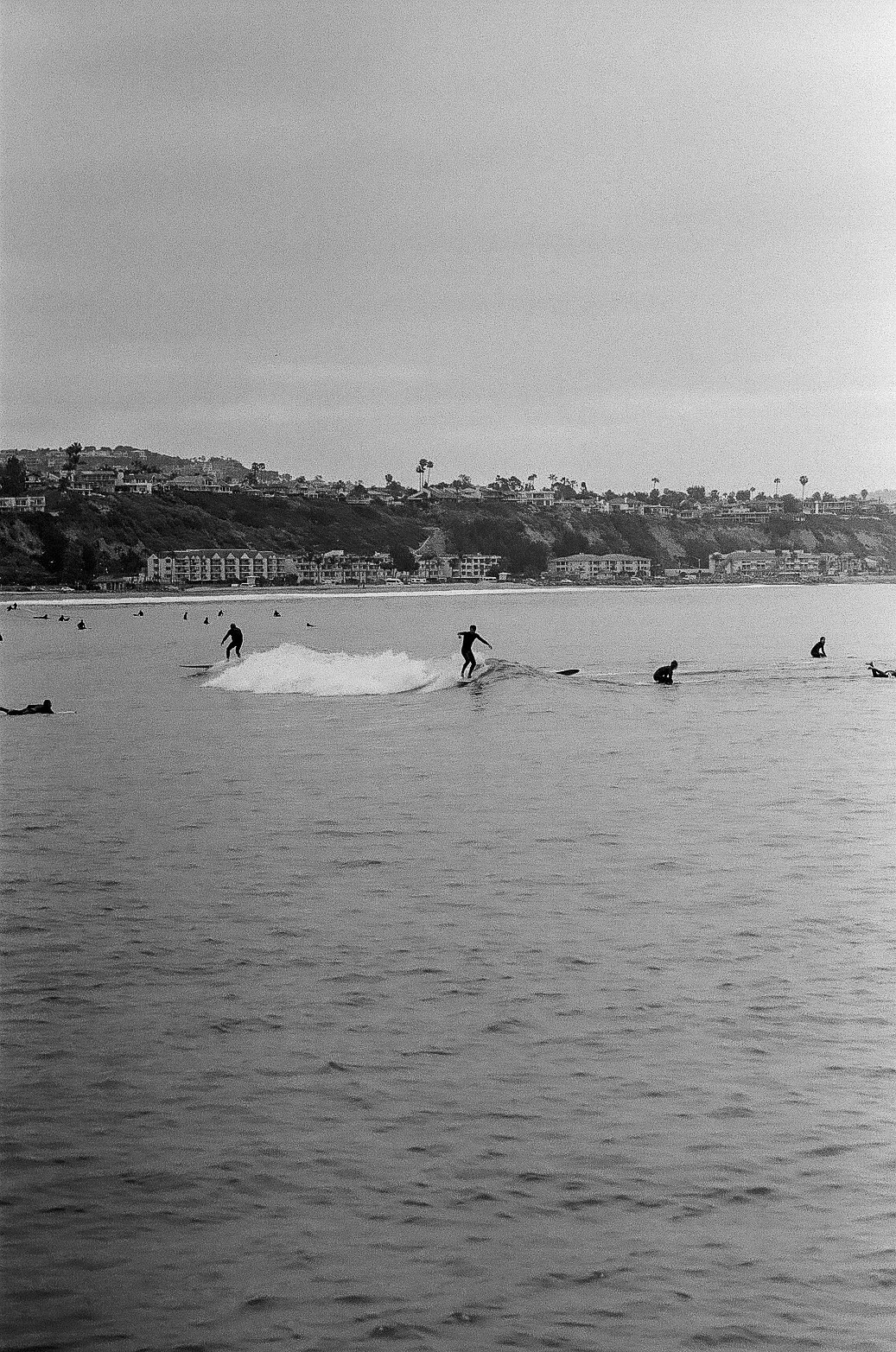 A black and white photo of a beach scene featuring a surfer riding a wave with other surfers and swimmers in the water, and buildings with palm trees on a hillside in the background.