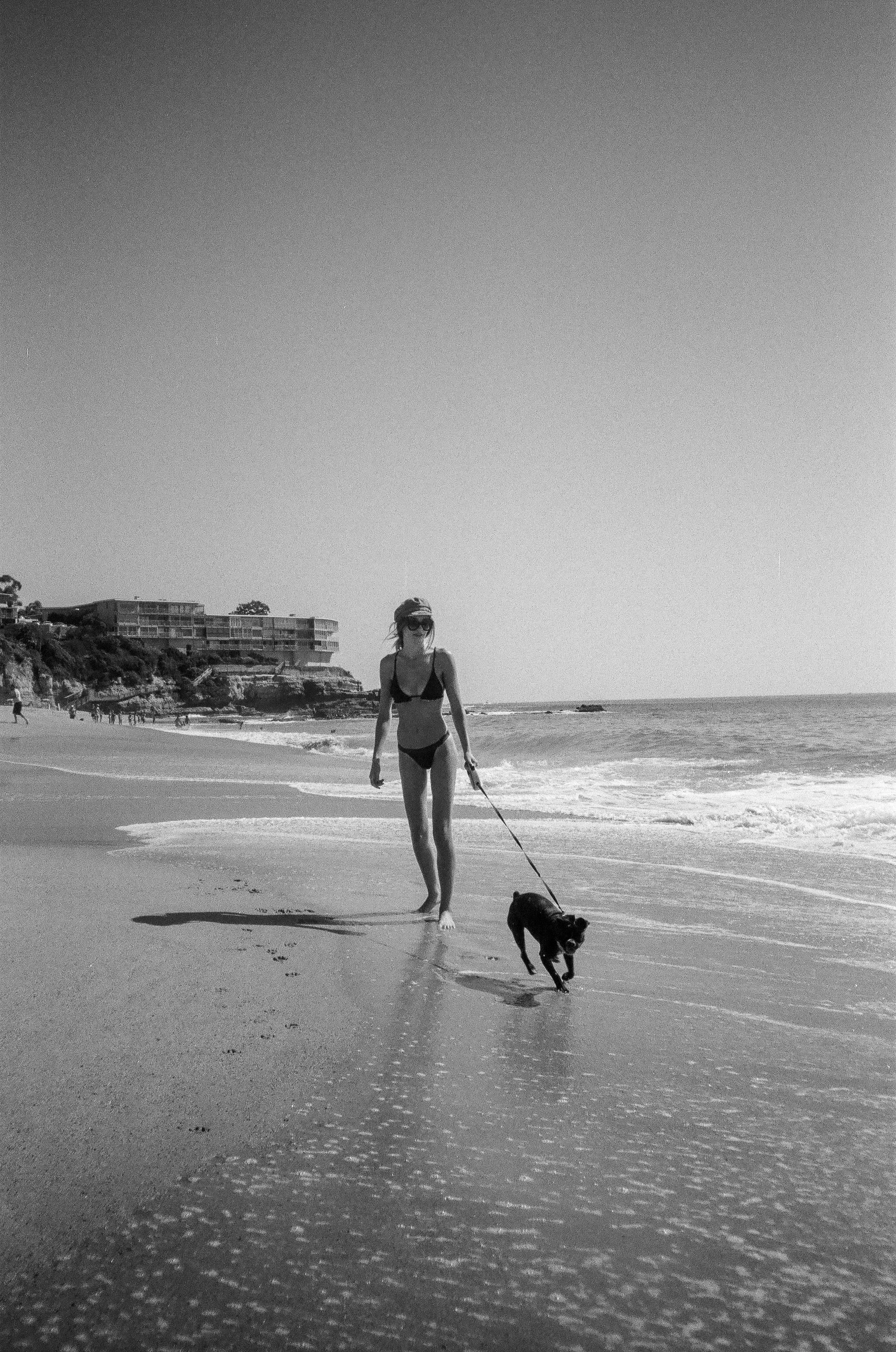 A woman walking her dog along the beach with buildings in the background, black and white photo.