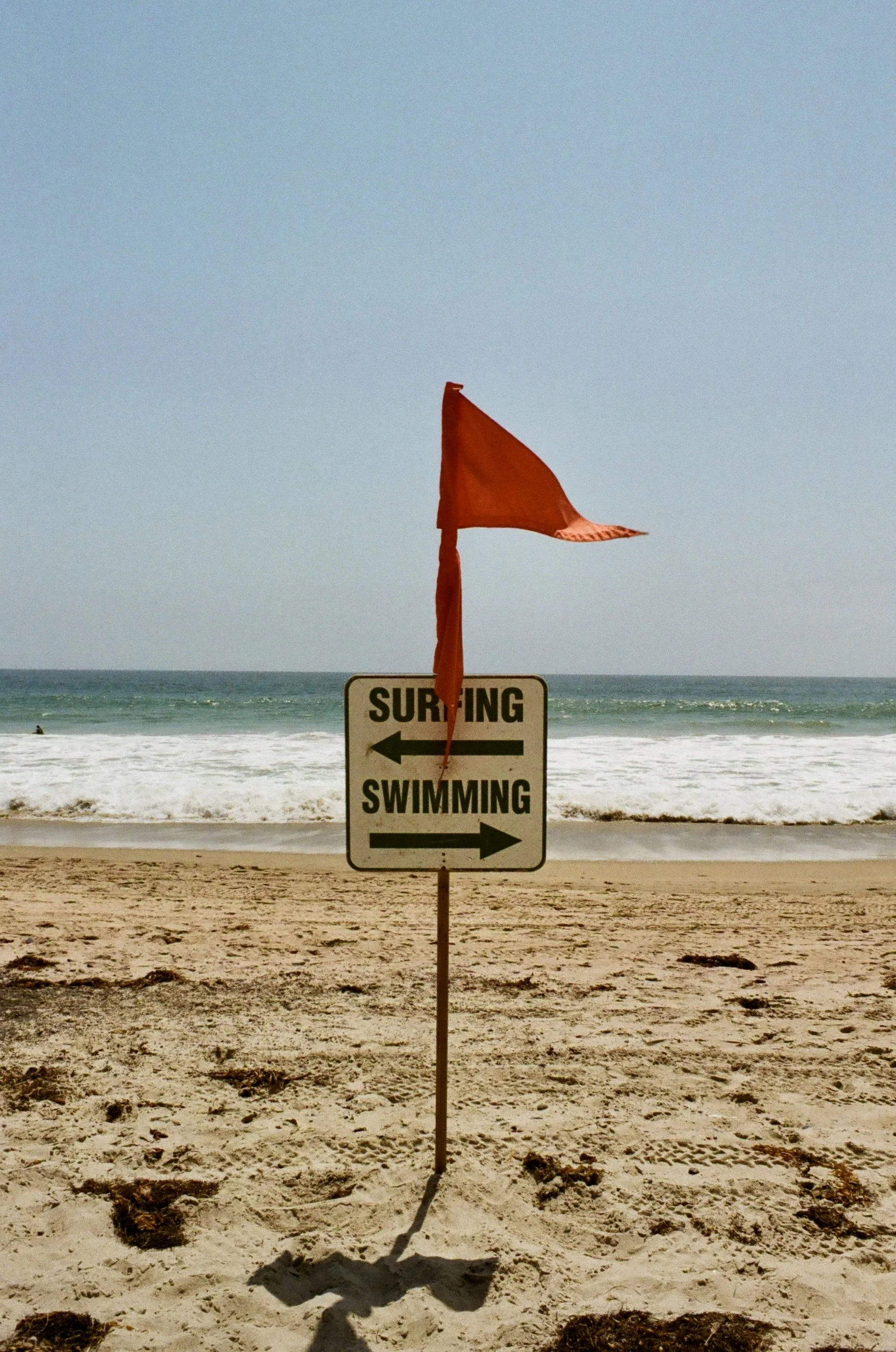 Beach scene with a sign indicating different directions for surfing and swimming, a red flag, and the ocean in the background.