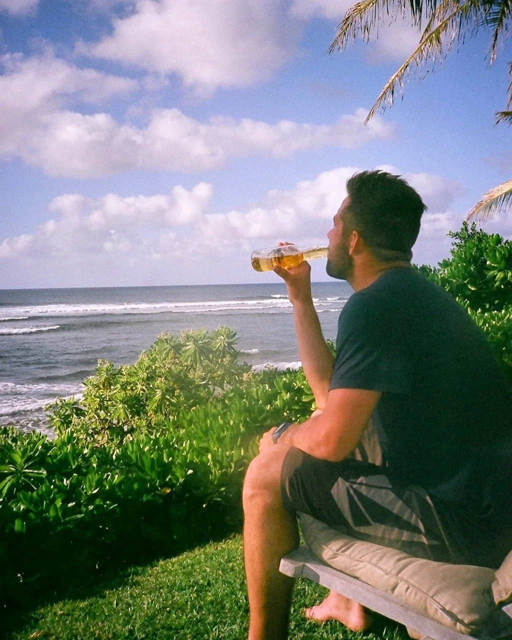 A man sitting on a wooden bench by the beach, drinking a bottle of beer while facing the ocean, surrounded by lush green plants and palm trees under a partly cloudy sky.