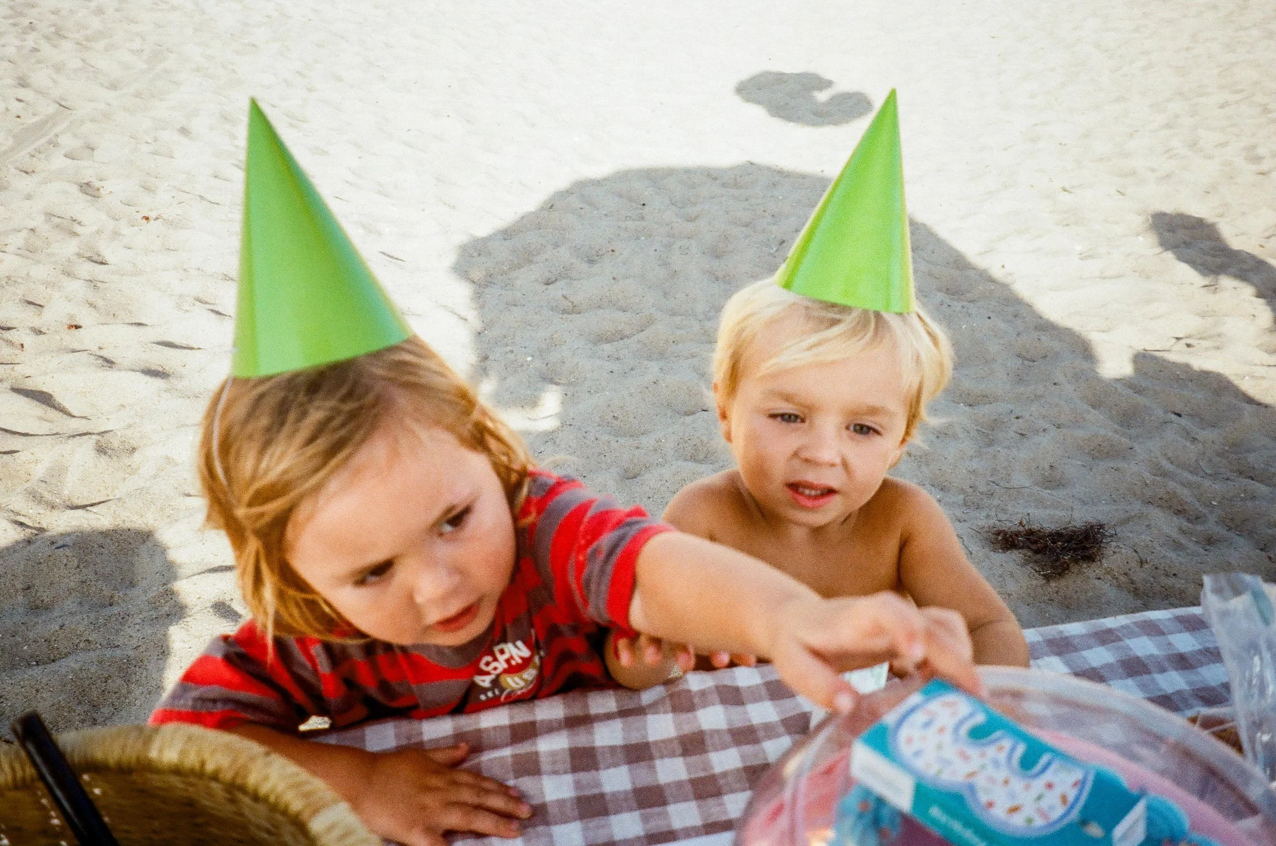 Two children wearing green party hats sitting at a picnic table on a beach, reaching for party favors and looking curious.
